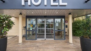 Entrada de hotel con puerta de vidrio, un letrero encima y plantas en macetas a ambos lados.
