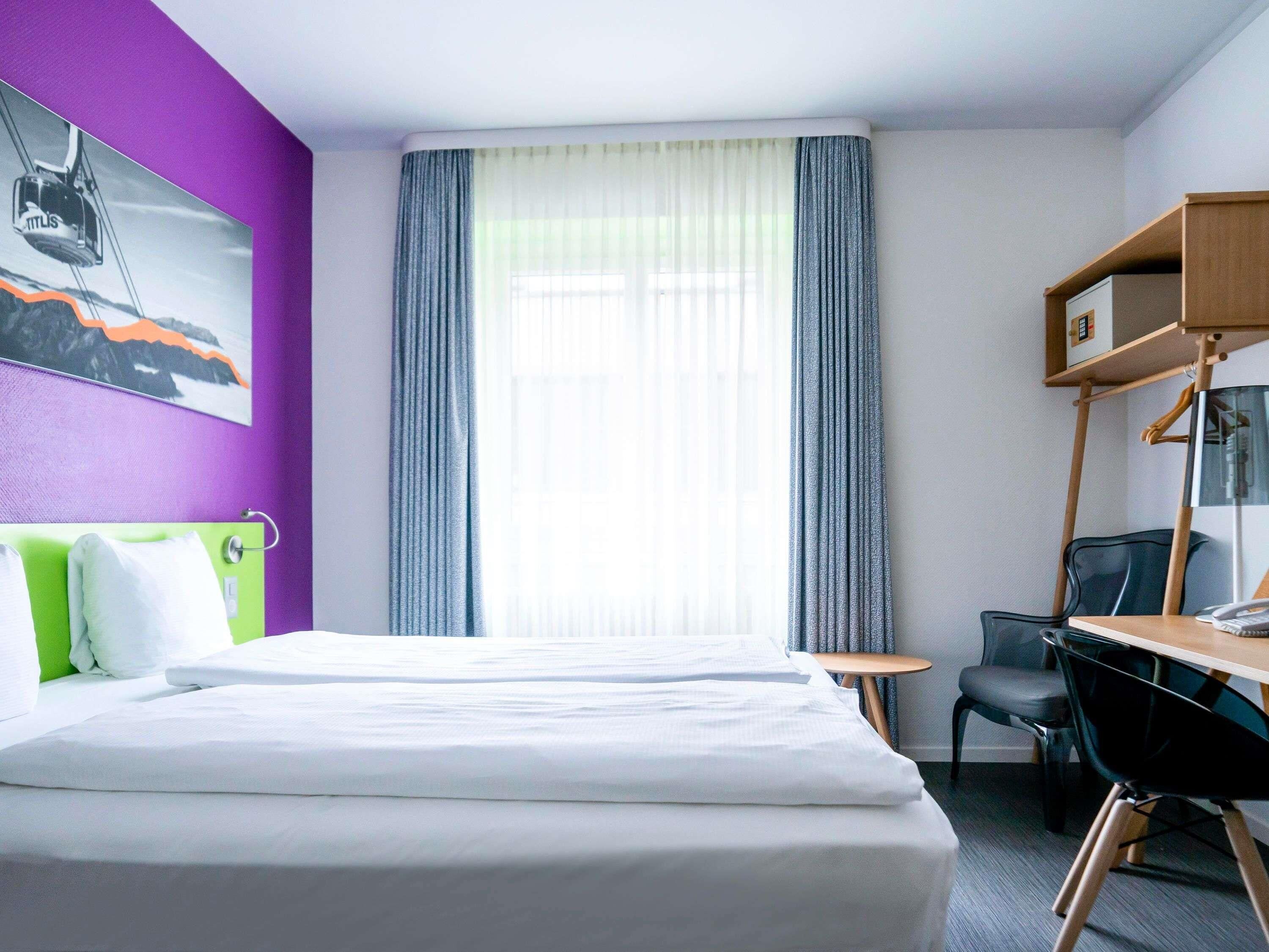 Una habitación de hotel con cama, escritorio, silla, una mesita y ventana con cortinas.