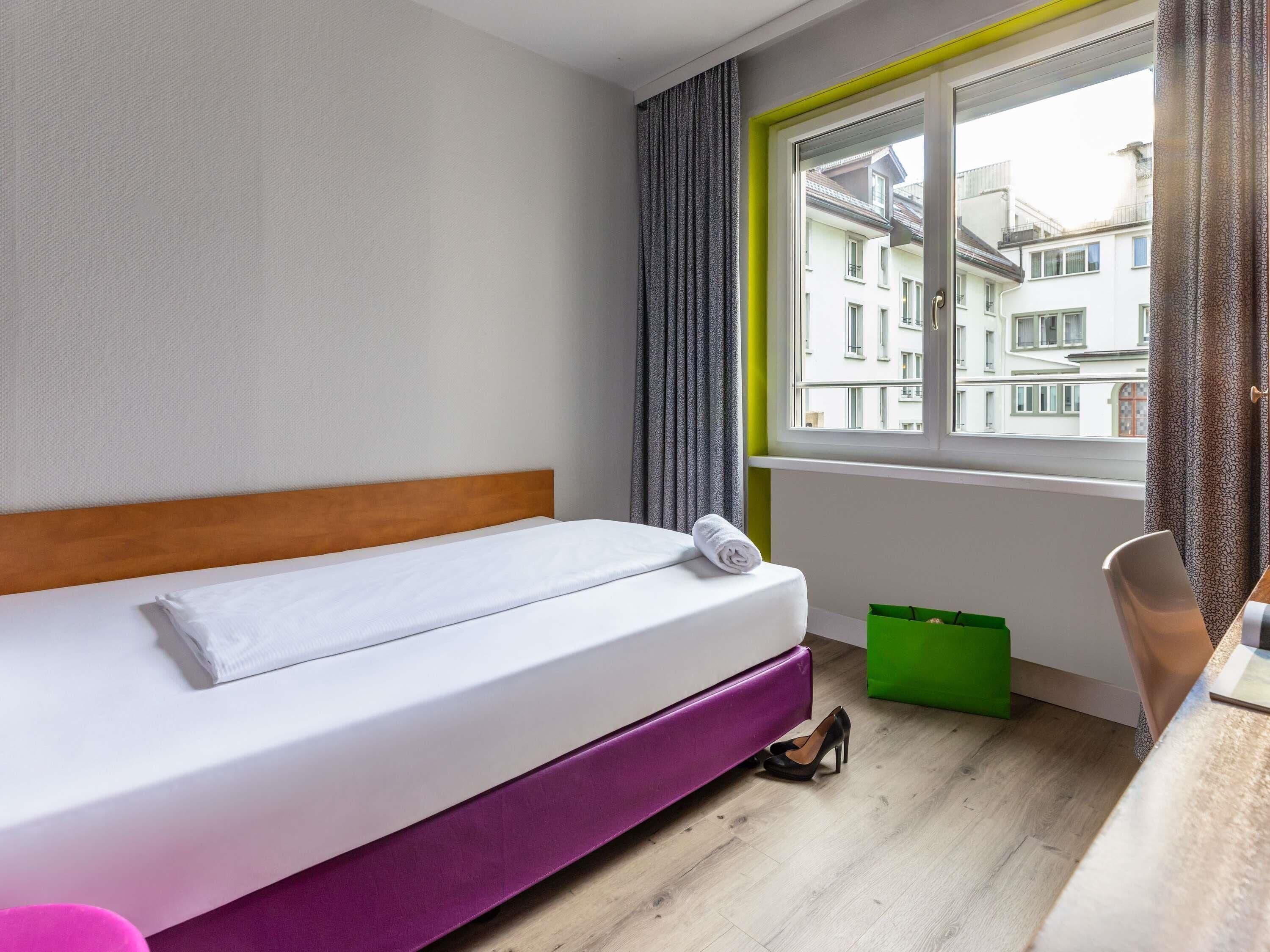A single bed with a wooden headboard, a window with curtains, a green storage box, and a chair.