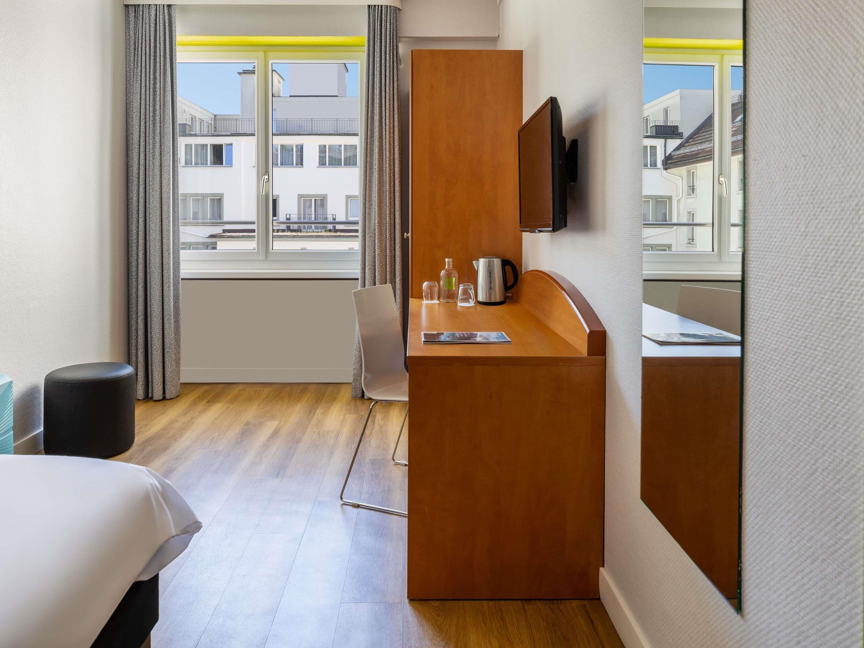 Hotellihuone, jossa on puinen työpöytä, tuoli, sänky, seinälle asennettu televisio ja ikkuna, jossa on verhot.