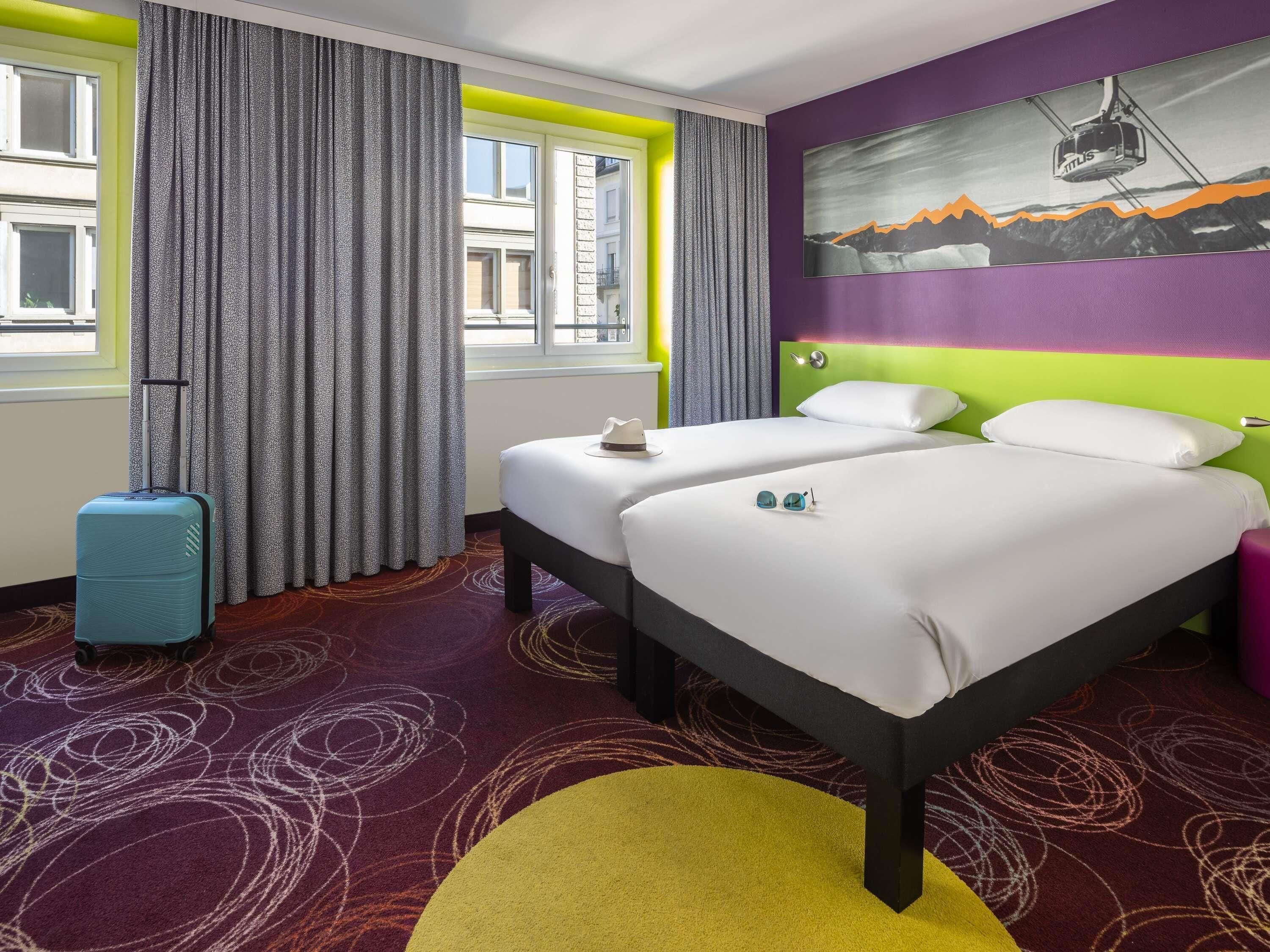 Habitación de hotel con dos camas, una ventana con cortinas, una maleta y un cuadro en la pared.