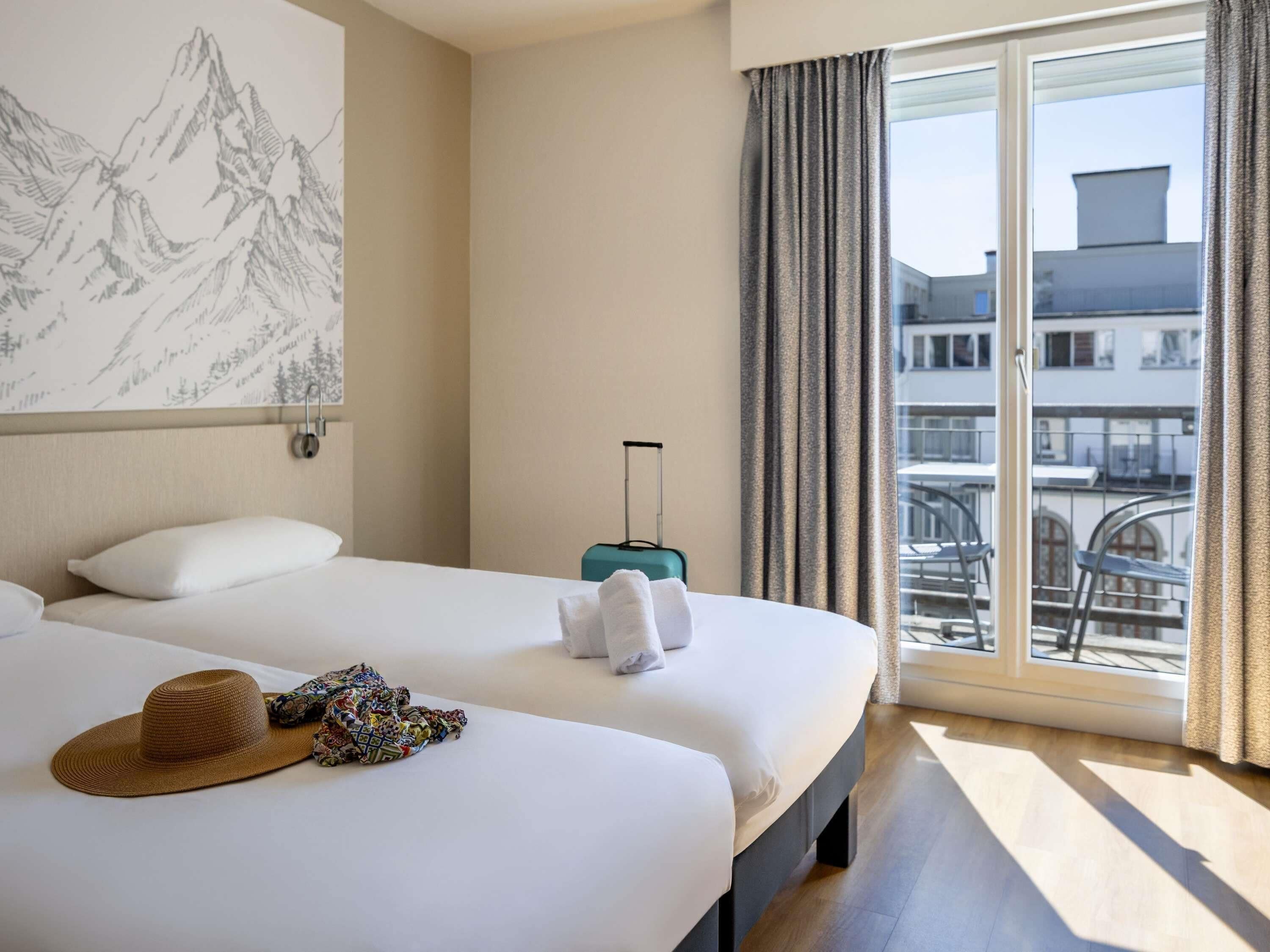 A hotel room with two beds, a large window with curtains, a suitcase, and a wall-mounted painting.