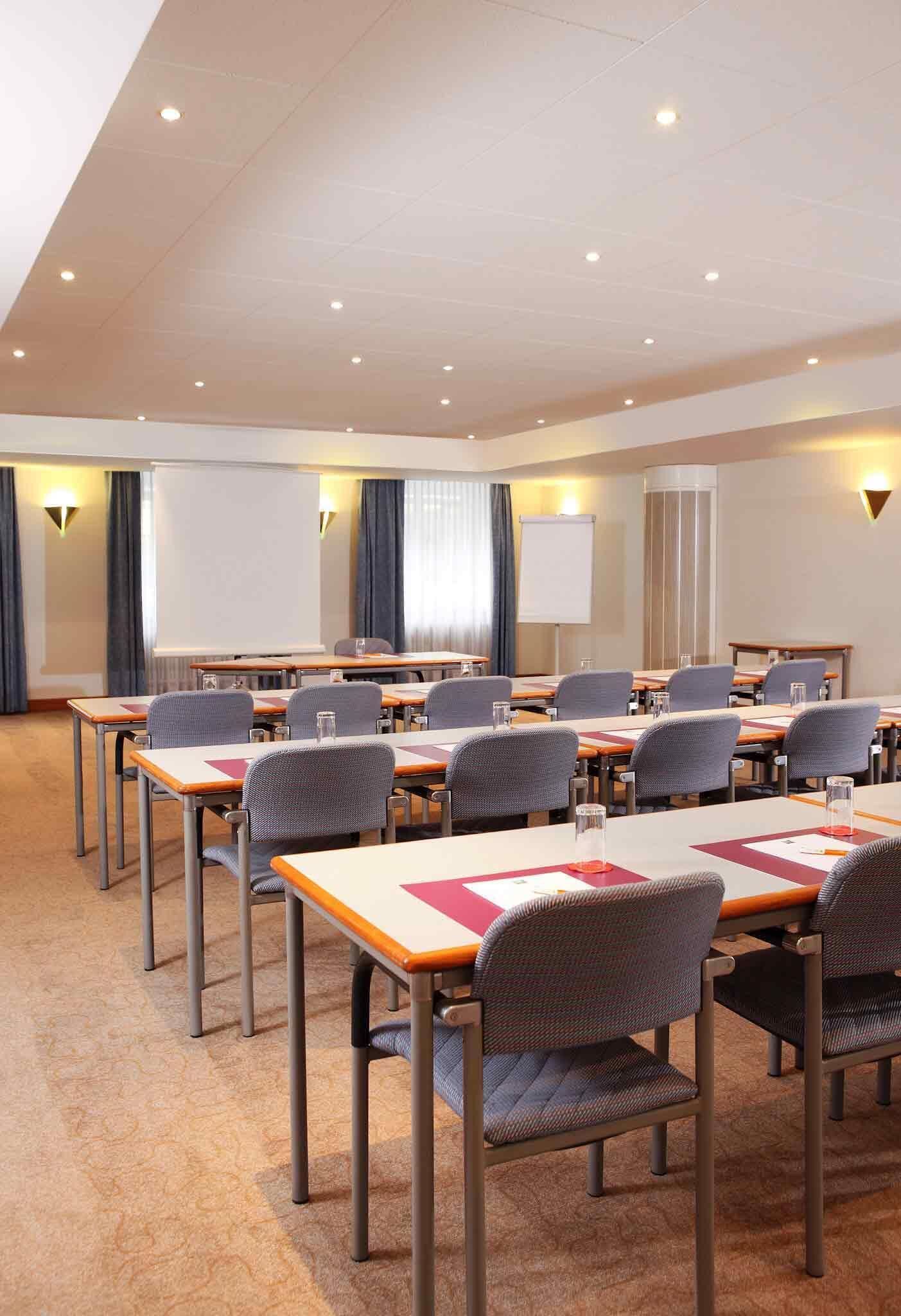 Un salón de conferencias con mesas, sillas, una pantalla de proyector y una pizarra.
