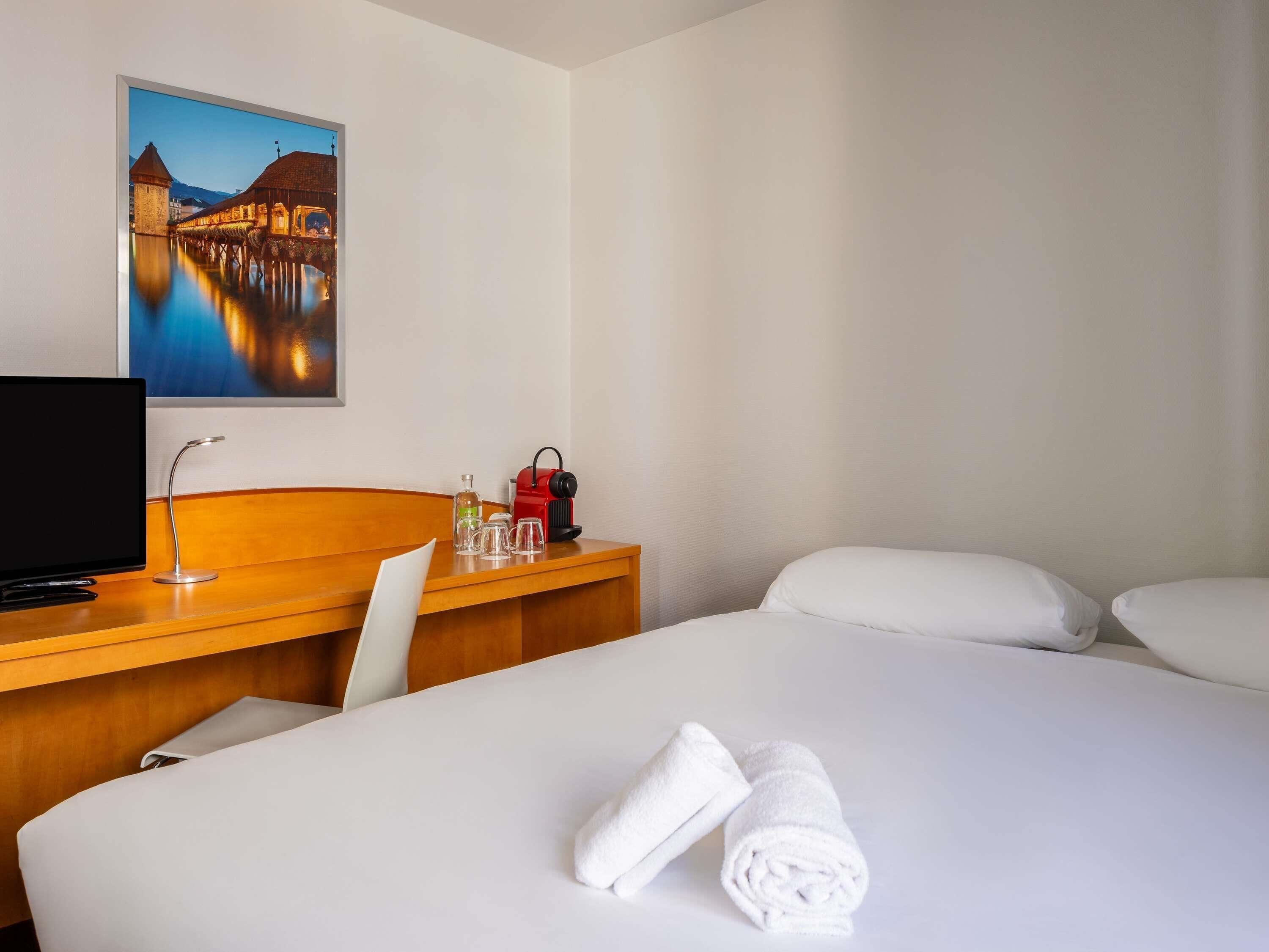 Habitación de hotel con una cama, un escritorio con televisor, un cuadro de un puente y una ventana.