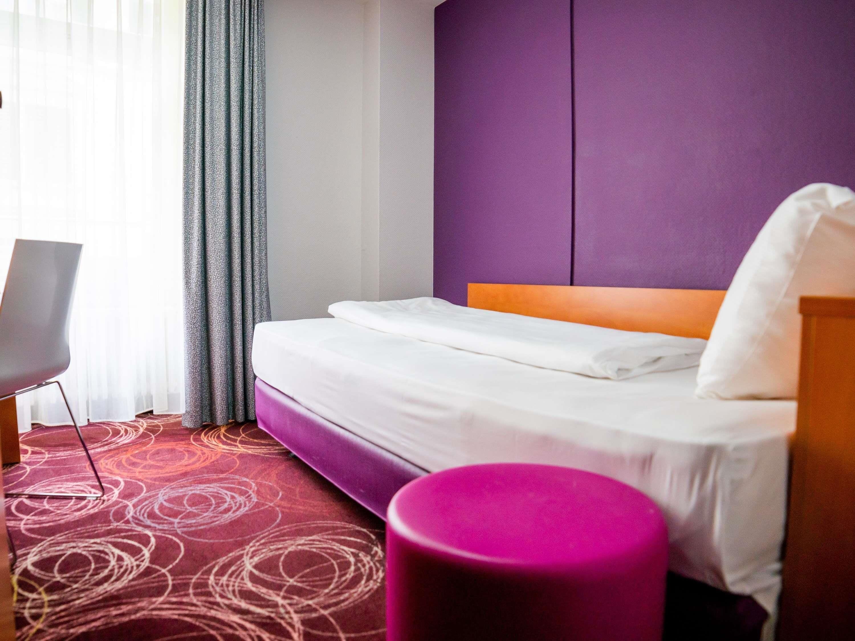 A hotel room with a bed, a chair, a purple ottoman, and a window with curtains.
