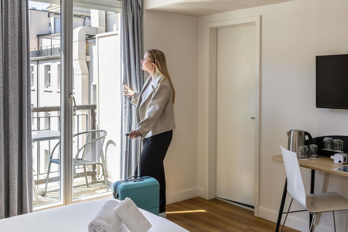 A hotel room with a bed, a suitcase, a desk with a kettle, and a chair.