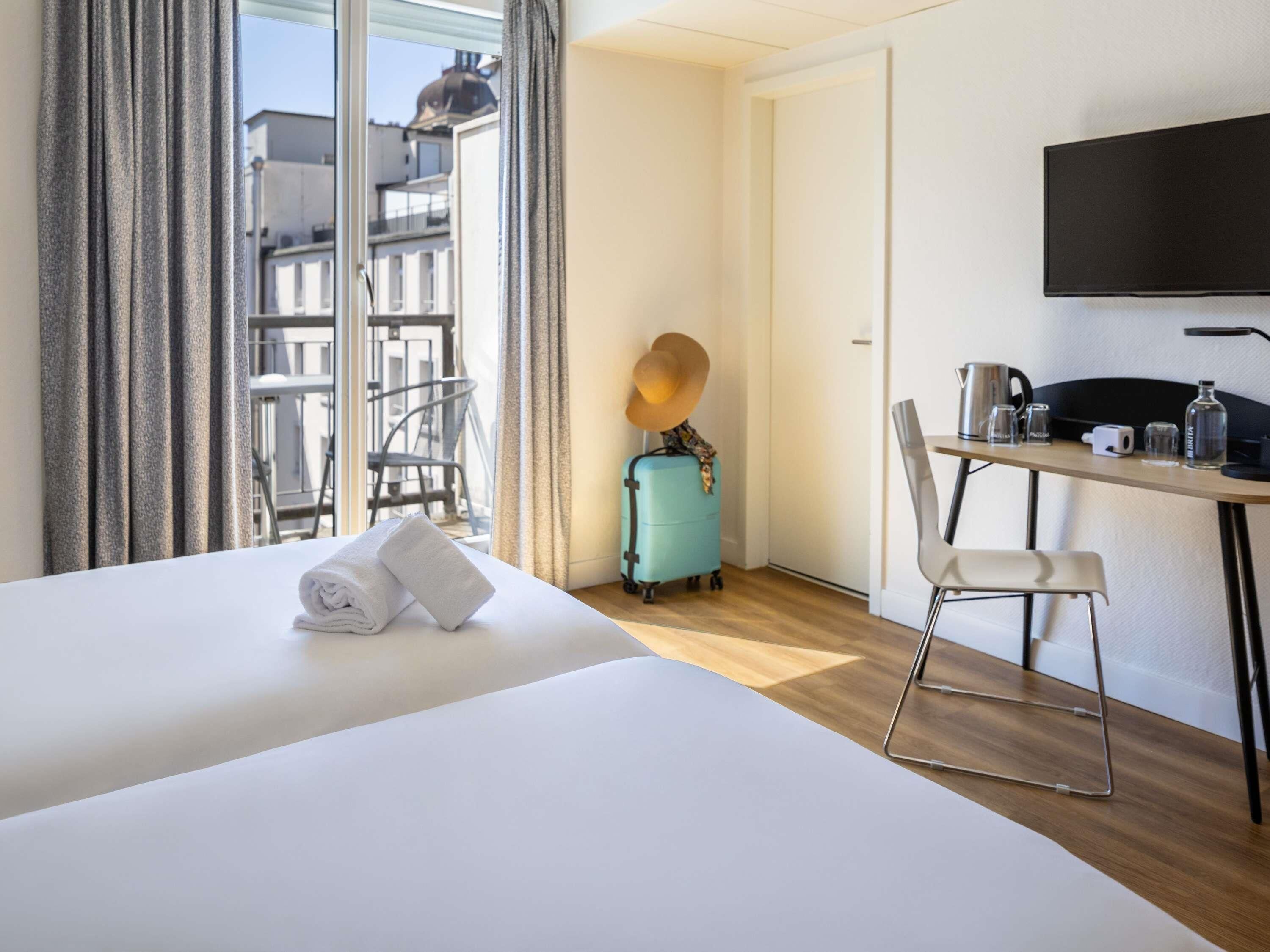 A hotel room with a bed, a desk with a chair, a suitcase, and a TV.