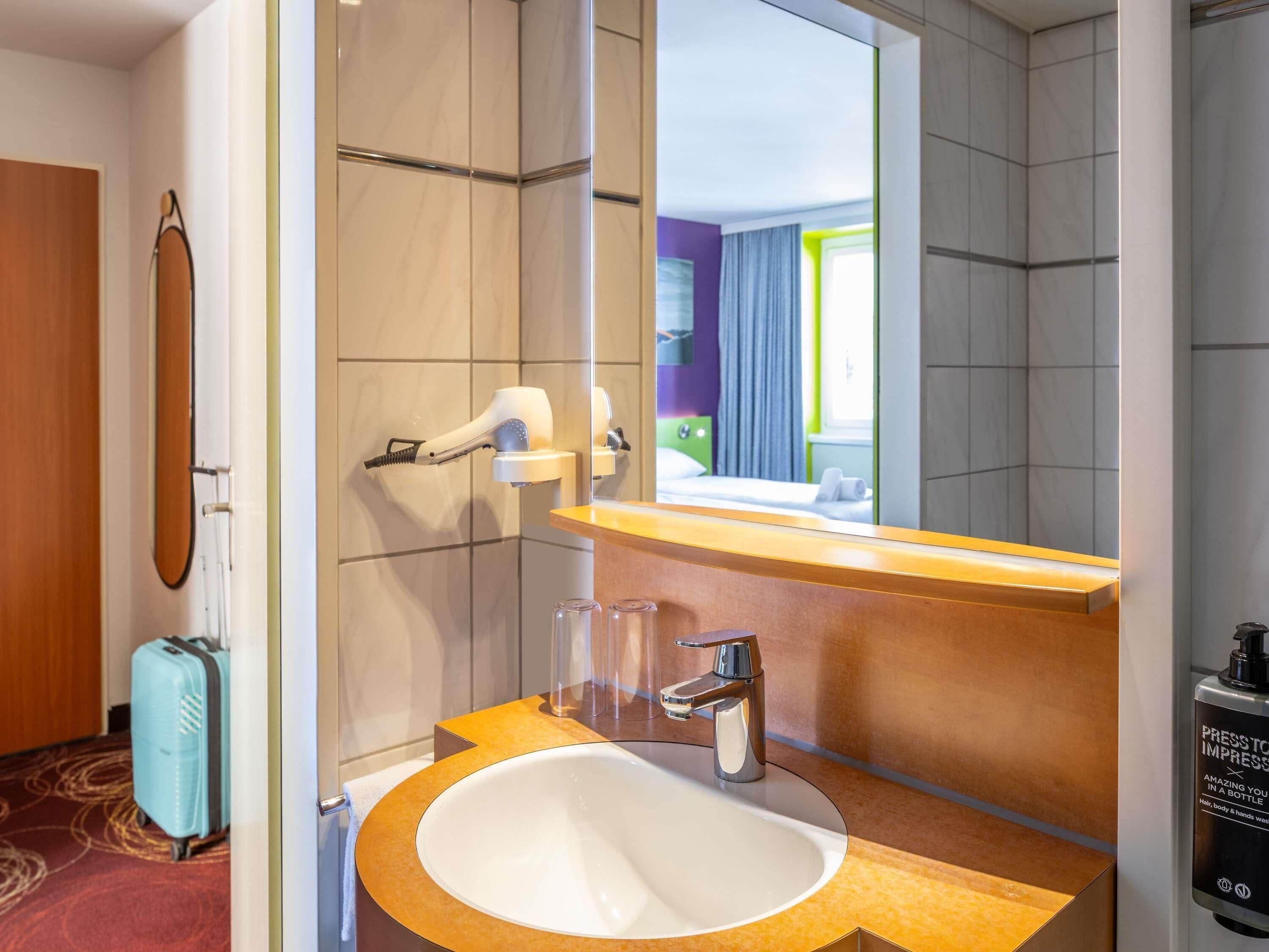 A hotel bathroom with a sink, mirror, towel rack, shower, and a suitcase.