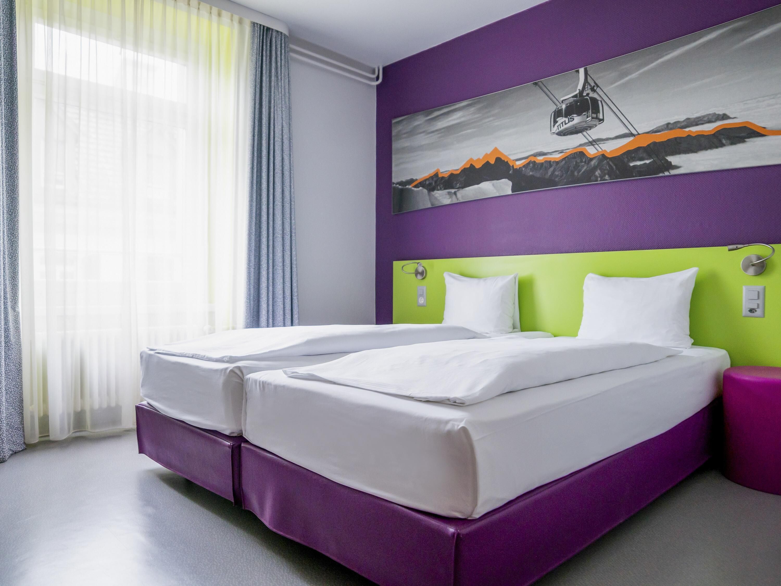 Ein Hotelzimmer mit zwei Betten, einem violetten Kopfteil, einem Gemälde einer Berglandschaft und einem Fenster mit transparenten Vorhängen.