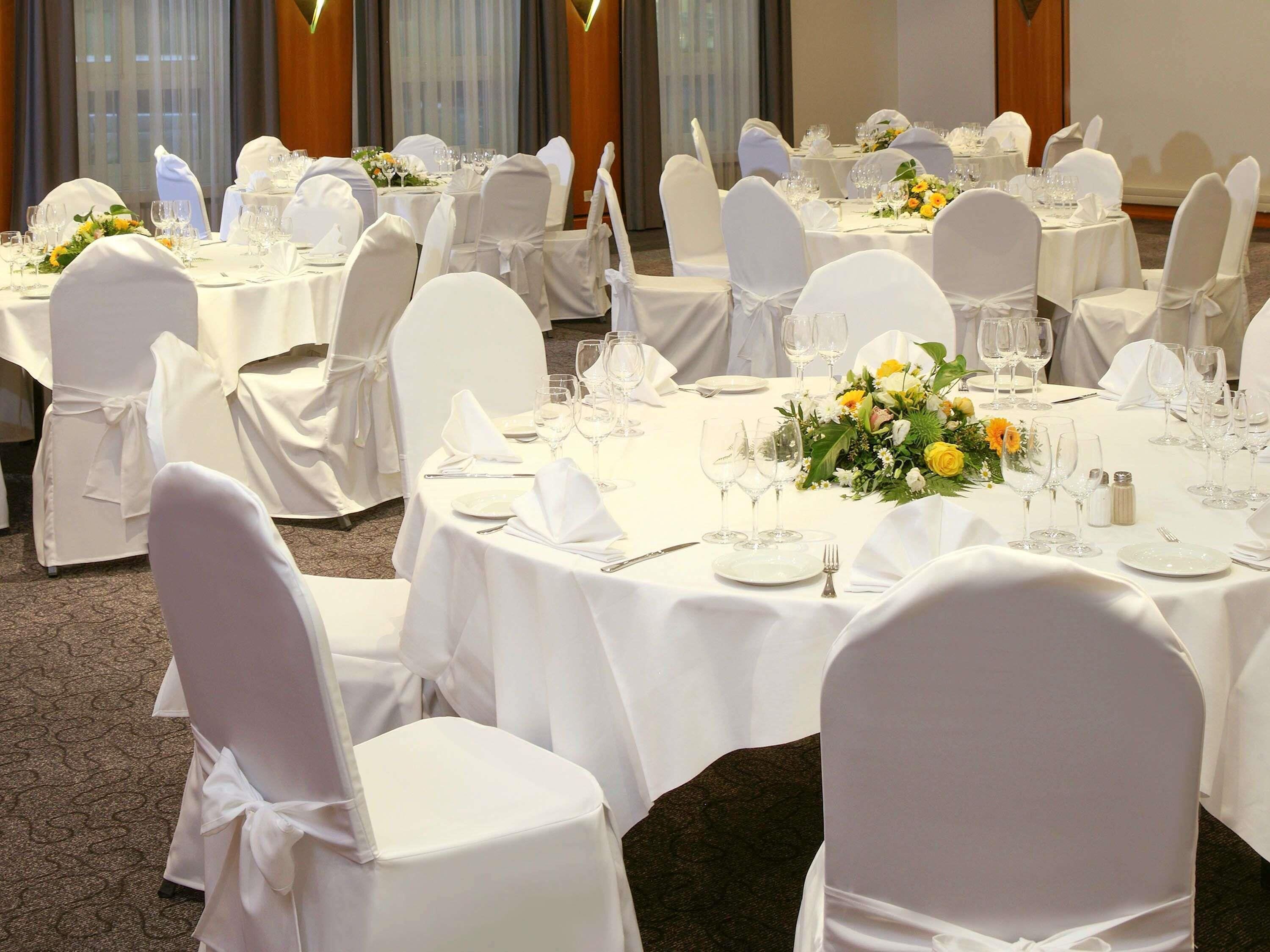 Un salón de banquetes con mesas redondas dispuestas para comer, sillas blancas y centros de mesa florales.