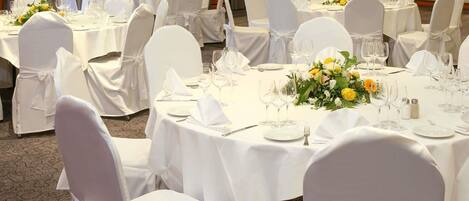 Un salón de banquetes con mesas redondas dispuestas para comer, sillas blancas y centros de mesa florales.