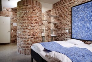 Un dormitorio con pared de ladrillo, cama con ropa de cama blanca, colcha azul estampada con flores y una gran obra de arte floral azul en la pared.