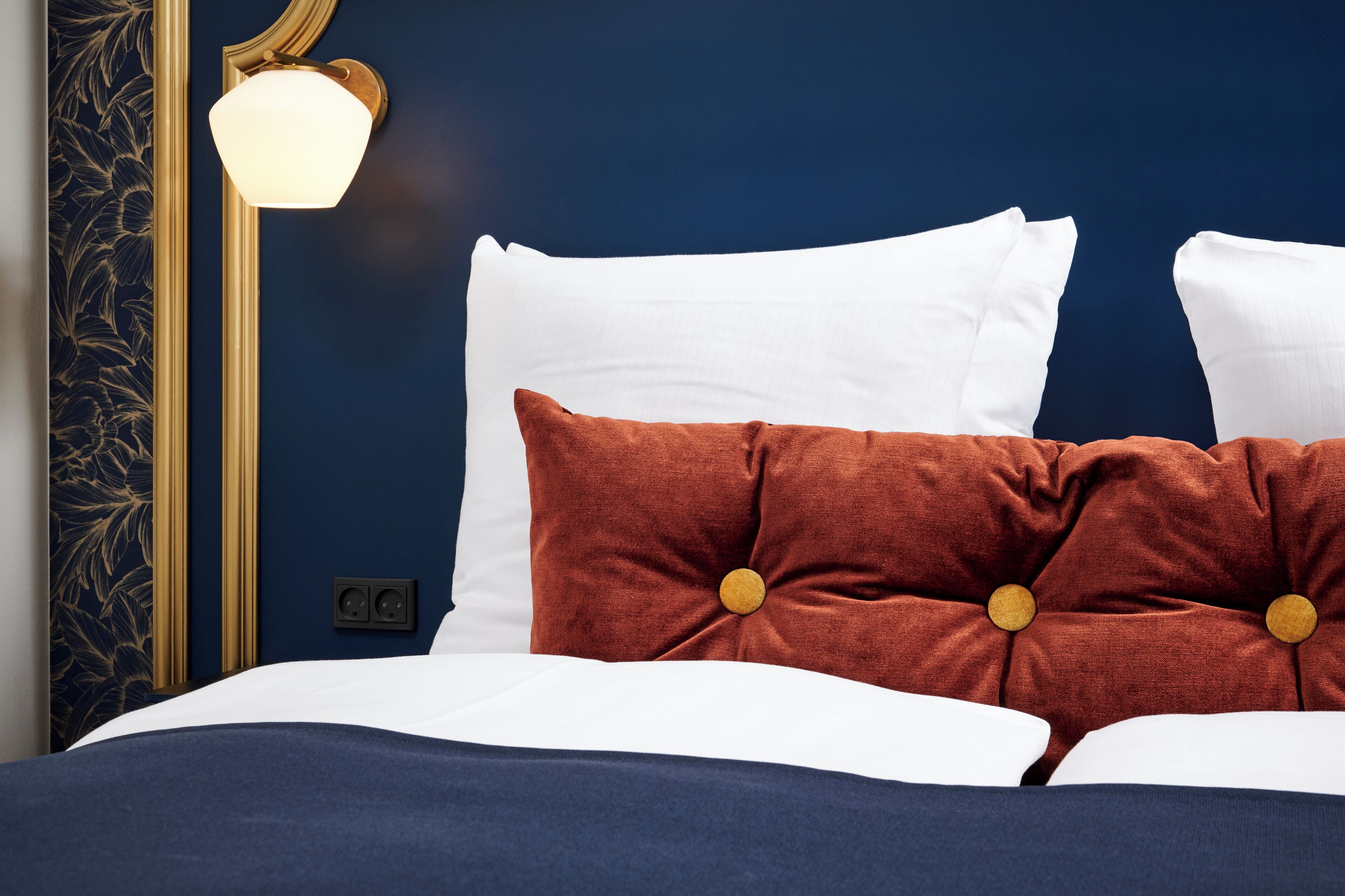 Una cama bien hecha con almohadas blancas y marrones, una pared azul oscuro y una lámpara de pared.