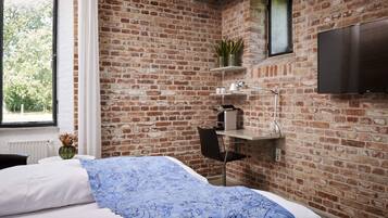 A bedroom with a brick wall, a window, a bed with a blue floral bedspread, a desk with a lamp, and a TV mounted on the wall.