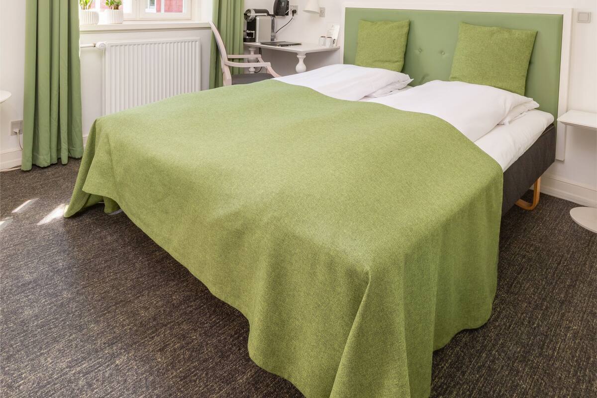 Habitación de hotel con cama, escritorio, silla, radiador, ventana con cortinas y un certificado enmarcado en la pared.