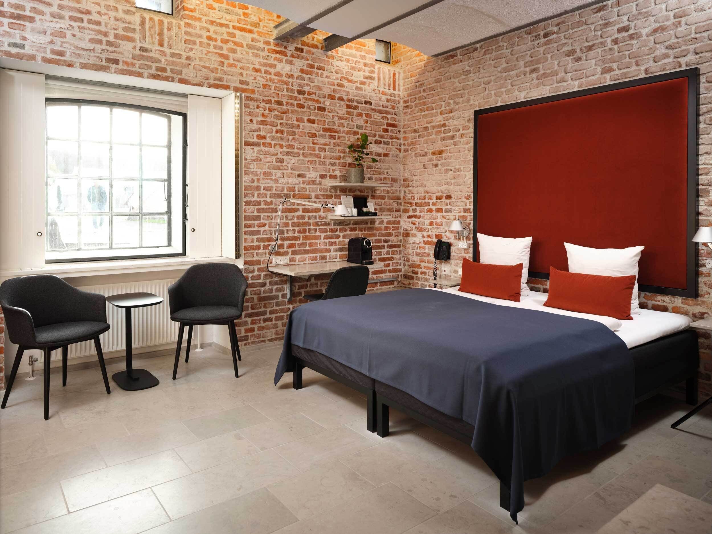 A hotel room with a brick wall, a large window, a bed with red and white bedding, a desk with a chair, and a small table.