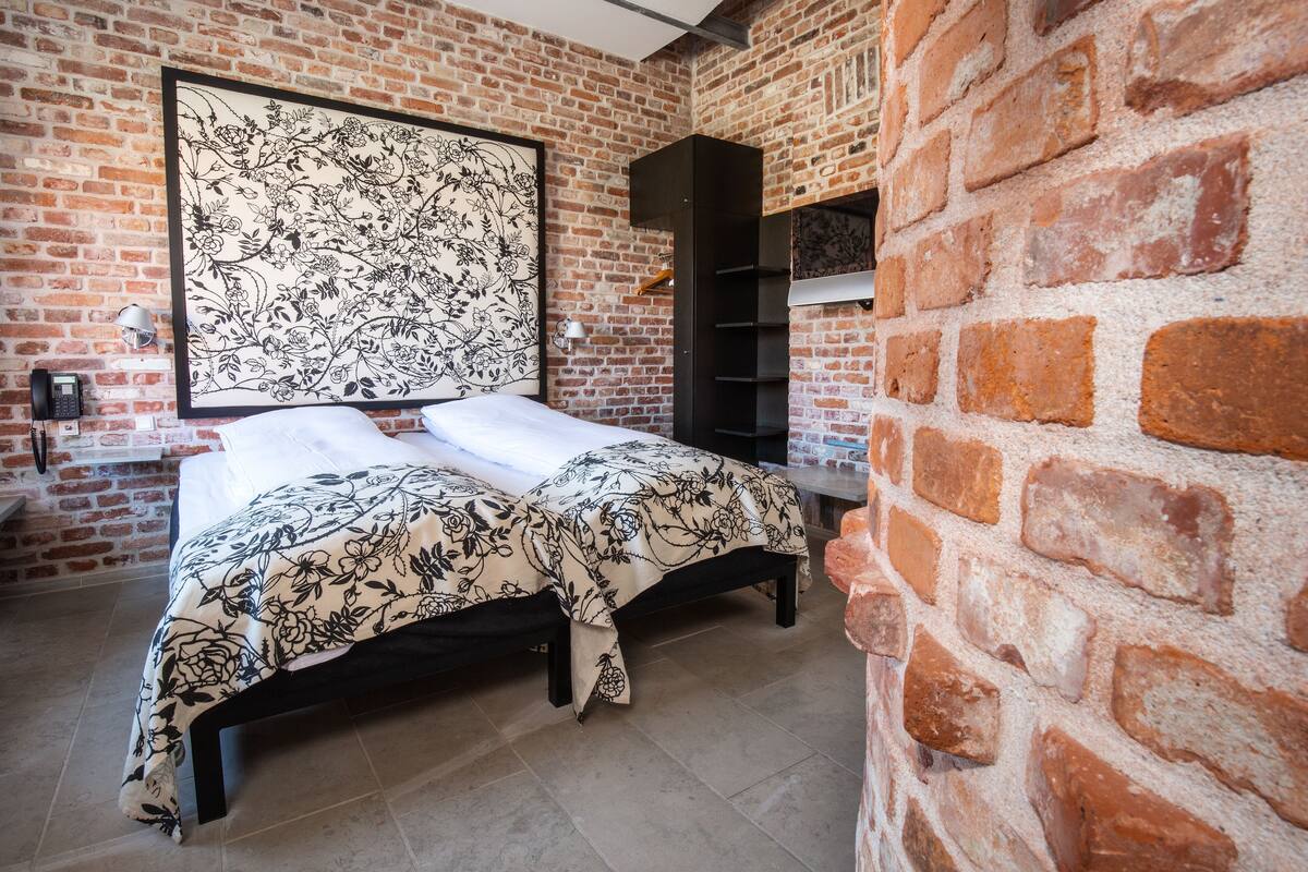 Una habitación con pared de ladrillo, una cama con cubrecama floral, cabecera negra y una estantería.