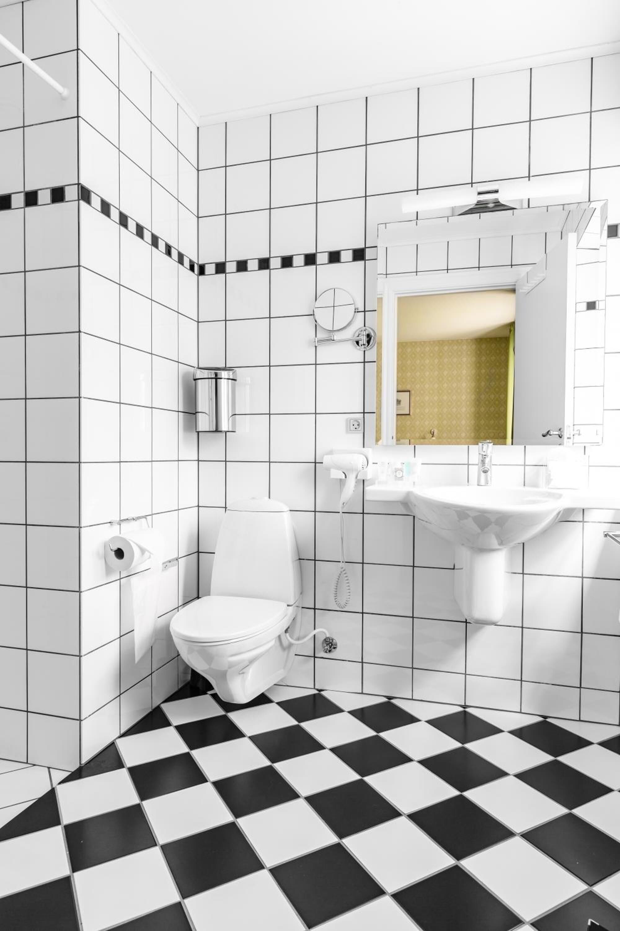 Un baño con azulejos blancos, piso a cuadros blanco y negro, inodoro blanco, lavamanos y espejo.