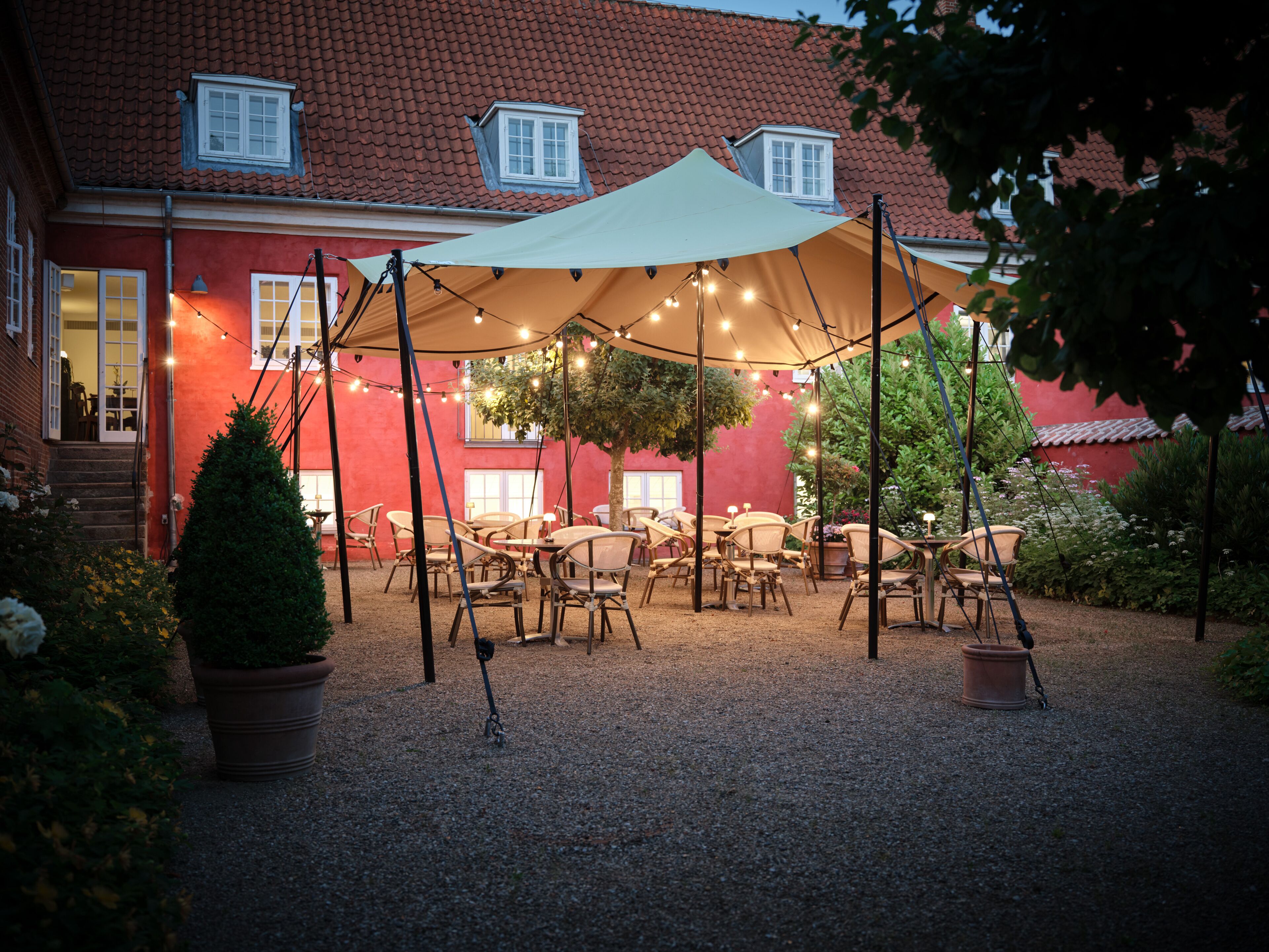 An outdoor seating area with tables and chairs under a canopy, surrounded by a gravel path and potted plants.