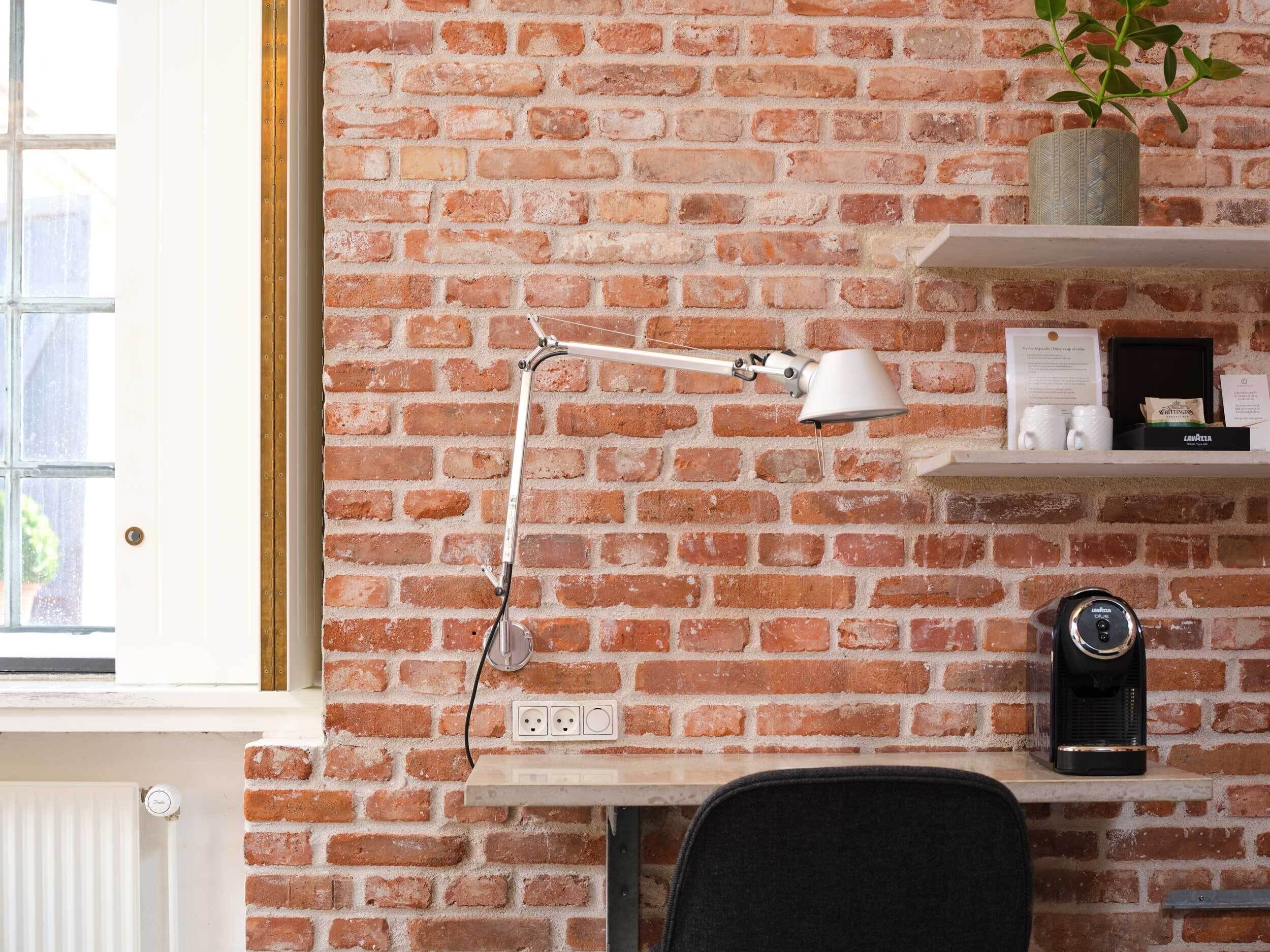 A room with a brick wall, a desk with a lamp, a coffee machine, and a potted plant.