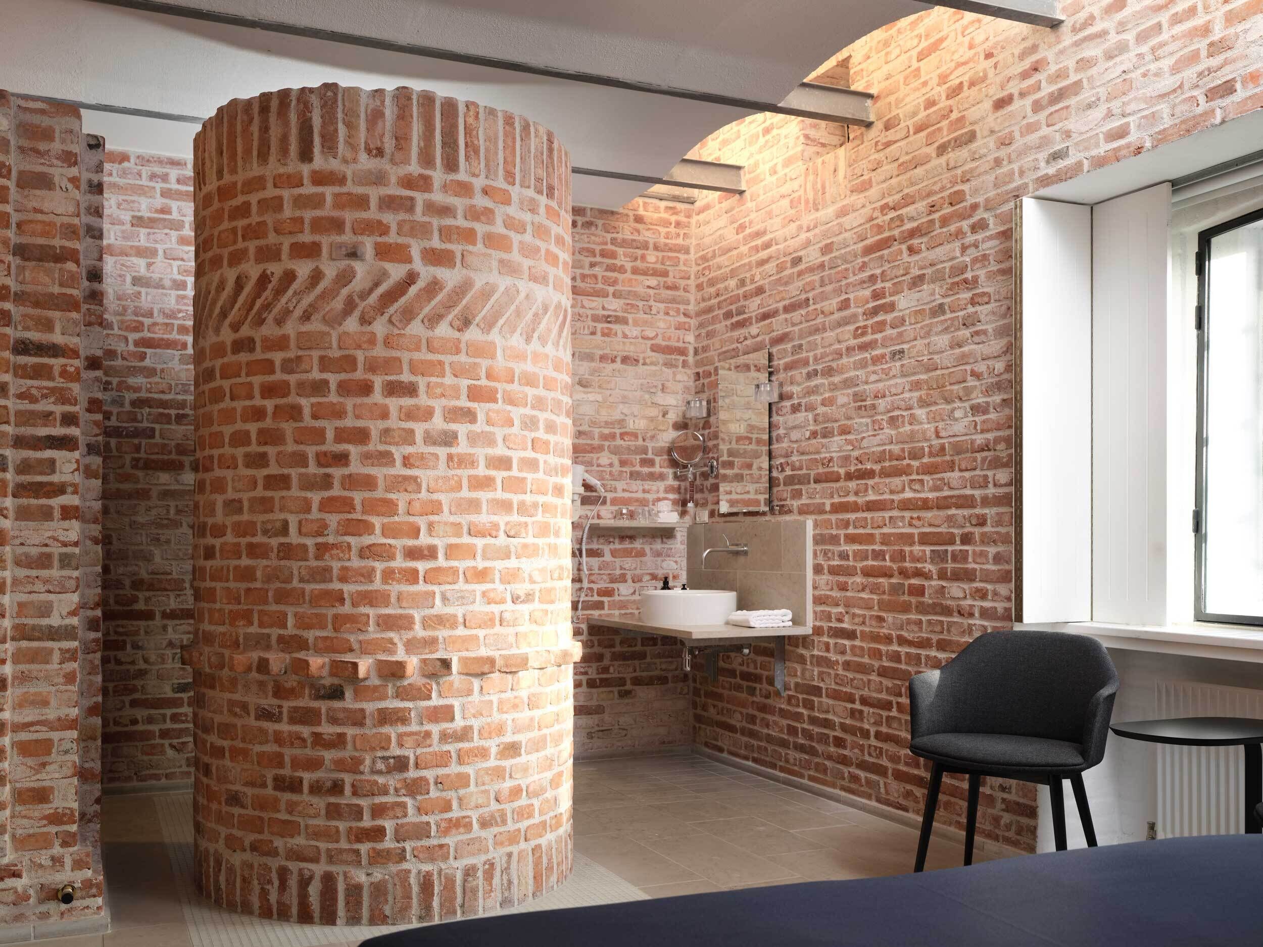 Un baño con pared de ladrillo, una columna circular, un lavabo, un espejo y una silla.