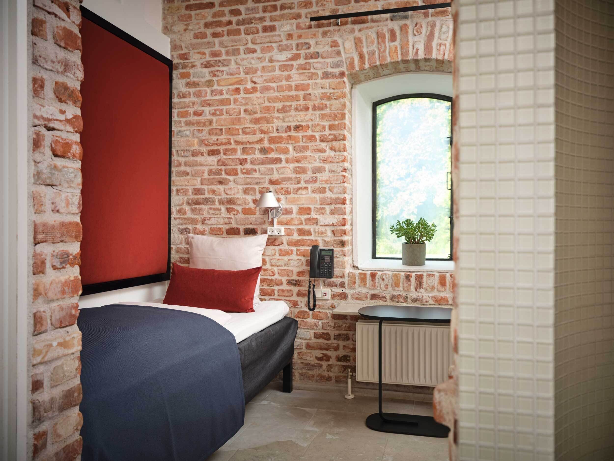 A bedroom with a brick wall, a window with a plant, a bedside table, and a bed with a red pillow.