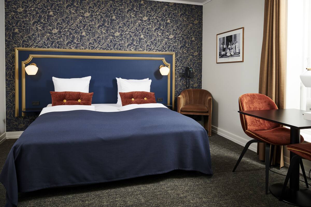 Habitación de hotel con una cama grande, dos almohadas, cabecera con apliques de pared, un sillón marrón, una mesa de madera y un cuadro enmarcado en la pared.