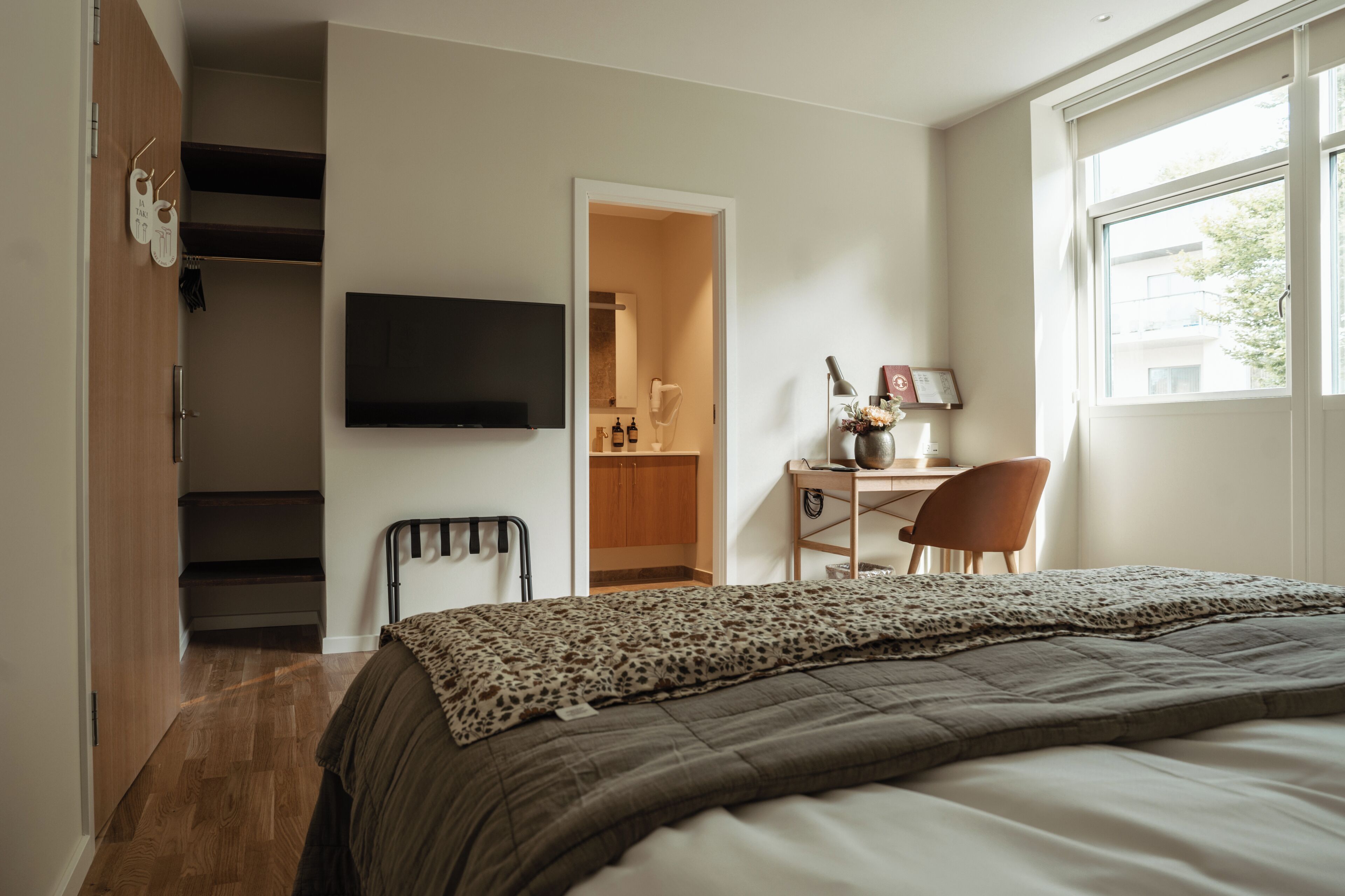 A bedroom with a bed, a desk, a chair, a TV, a wardrobe, and a window.