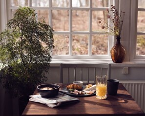 Una mesa de desayuno con periódico, un croissant y un vaso de jugo, una planta en maceta y un florero con flores secas cerca de una ventana.