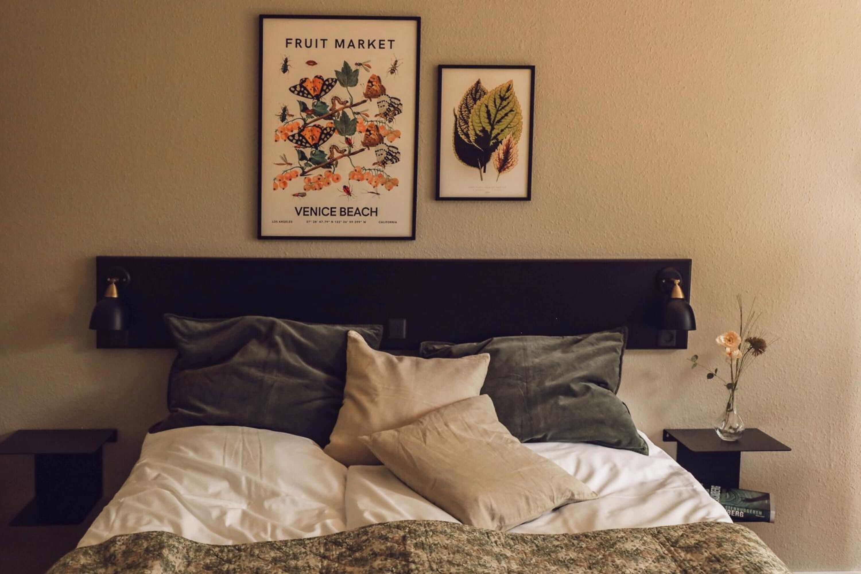 Una cama bien hecha con cabecera, dos almohadas y un cuadro con una imagen de un mercado de frutas.