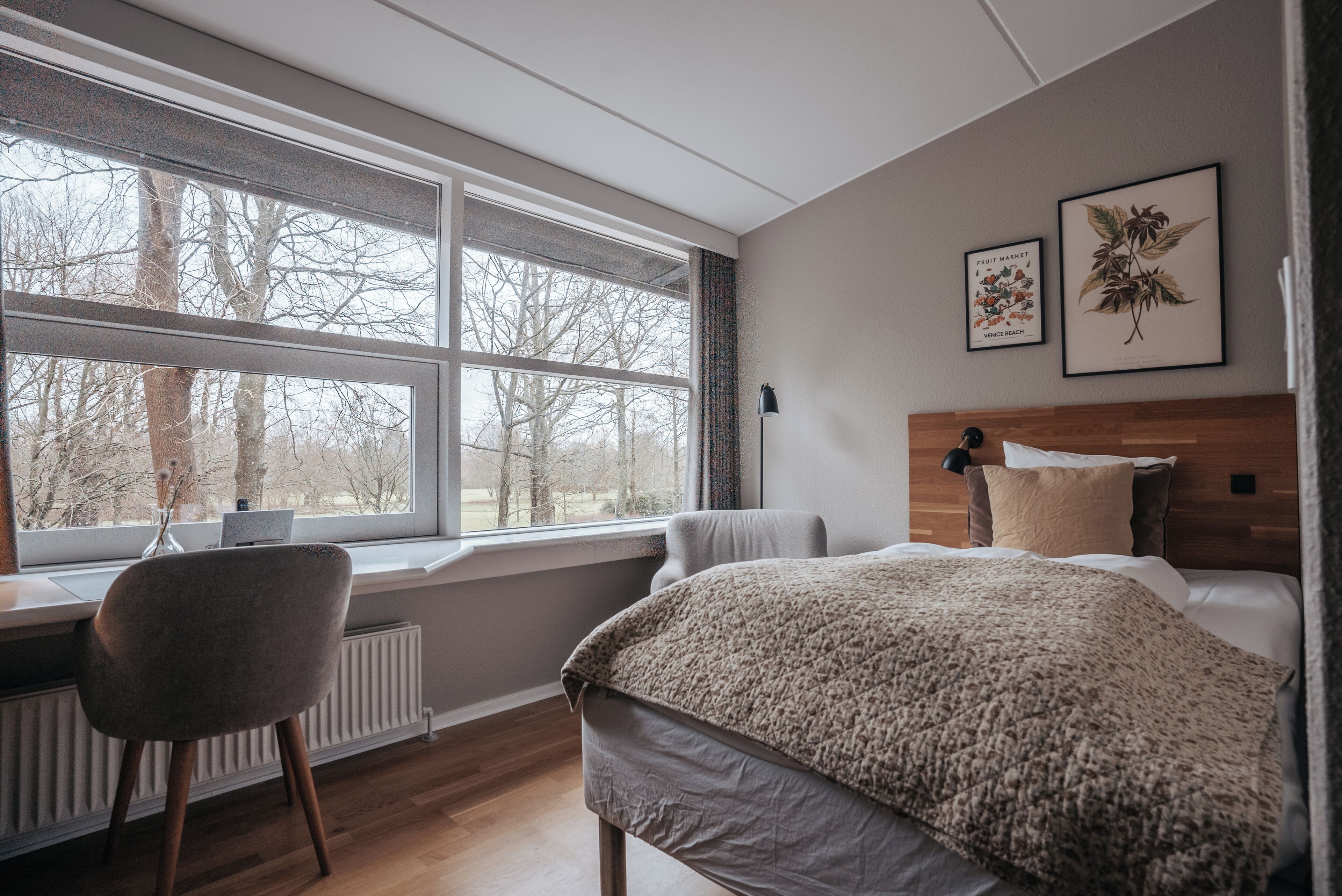 A bedroom with a large window, a bed, a chair, and two framed botanical prints on the wall.