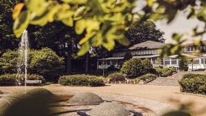 A garden with a fountain, a bench, and a building with a sloped roof. - Arnbjerg Pavillonen (Varde)