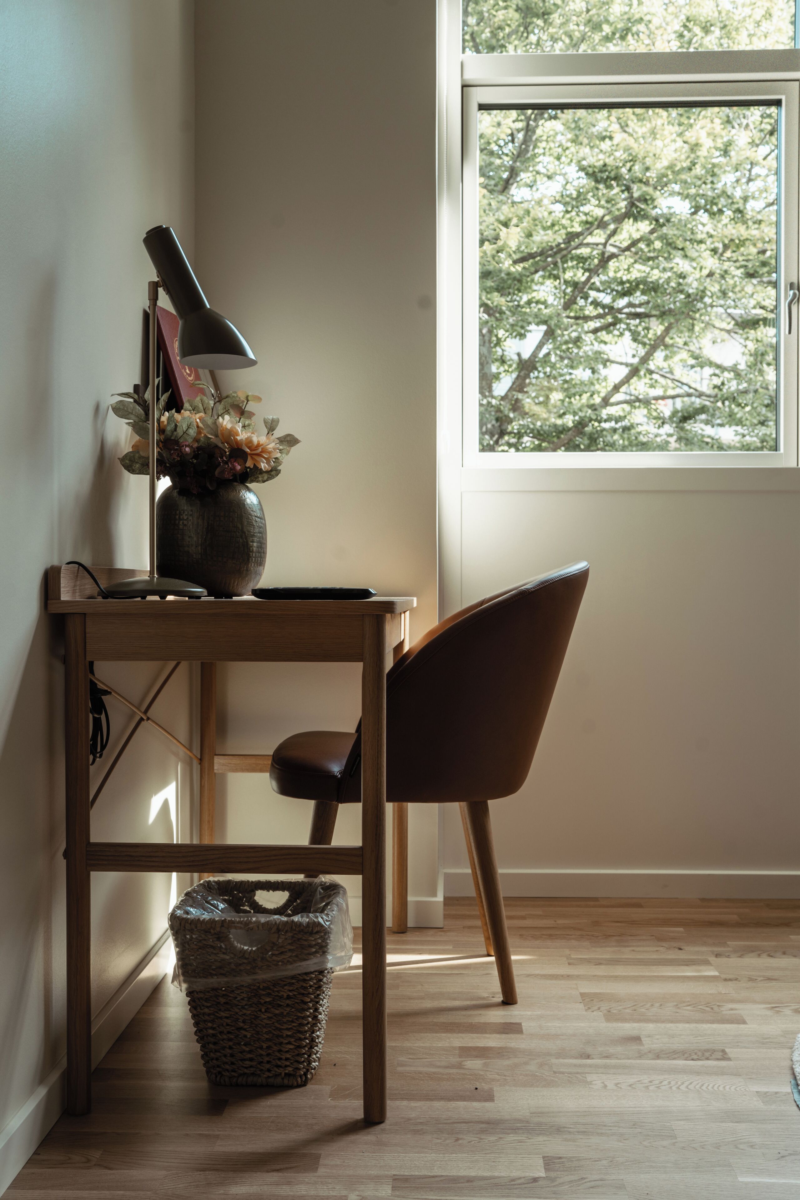 A wooden desk with a lamp, a vase with flowers, and a chair in a room with a window.