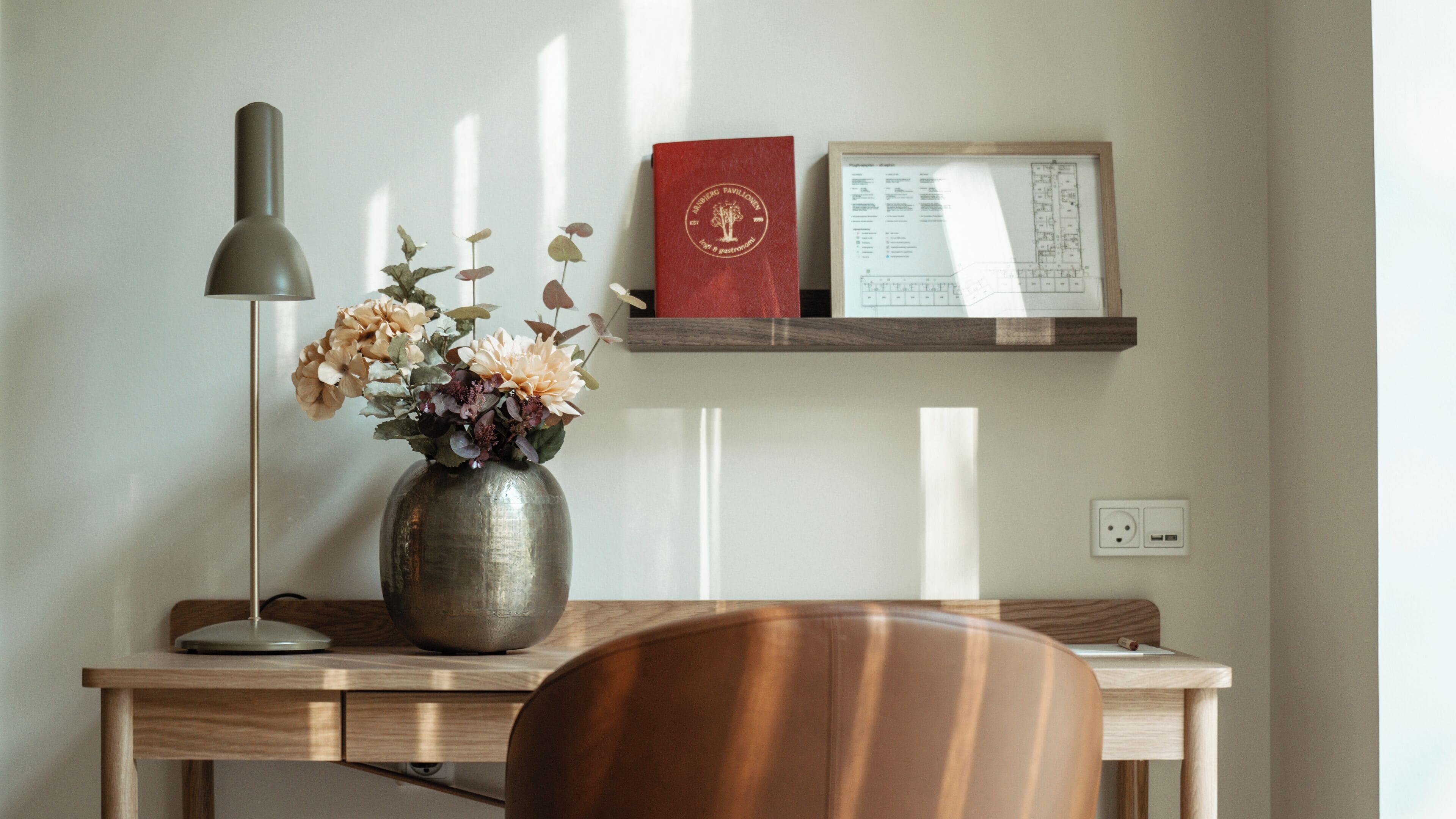 A desk with a vase of flowers, a lamp, a red book, and a framed architectural plan.