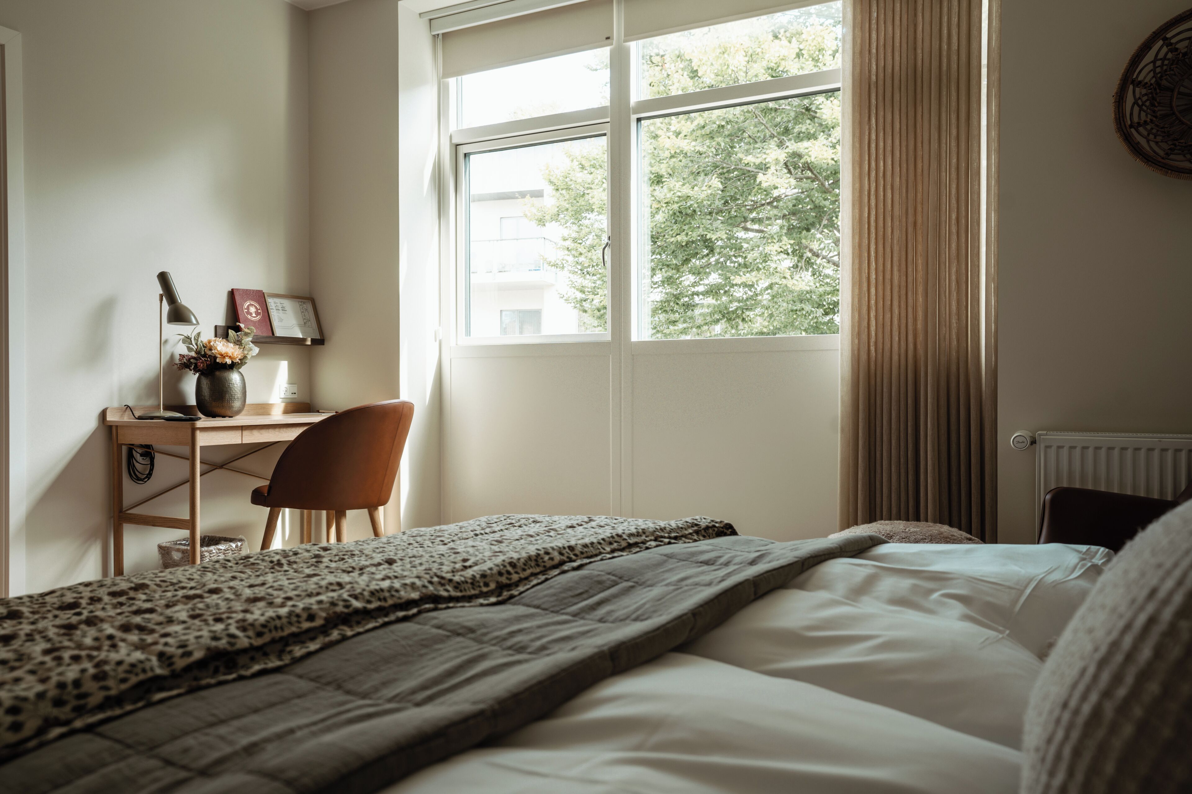 Une chambre à coucher avec un lit, un bureau et une chaise, une fenêtre avec des rideaux et un radiateur.