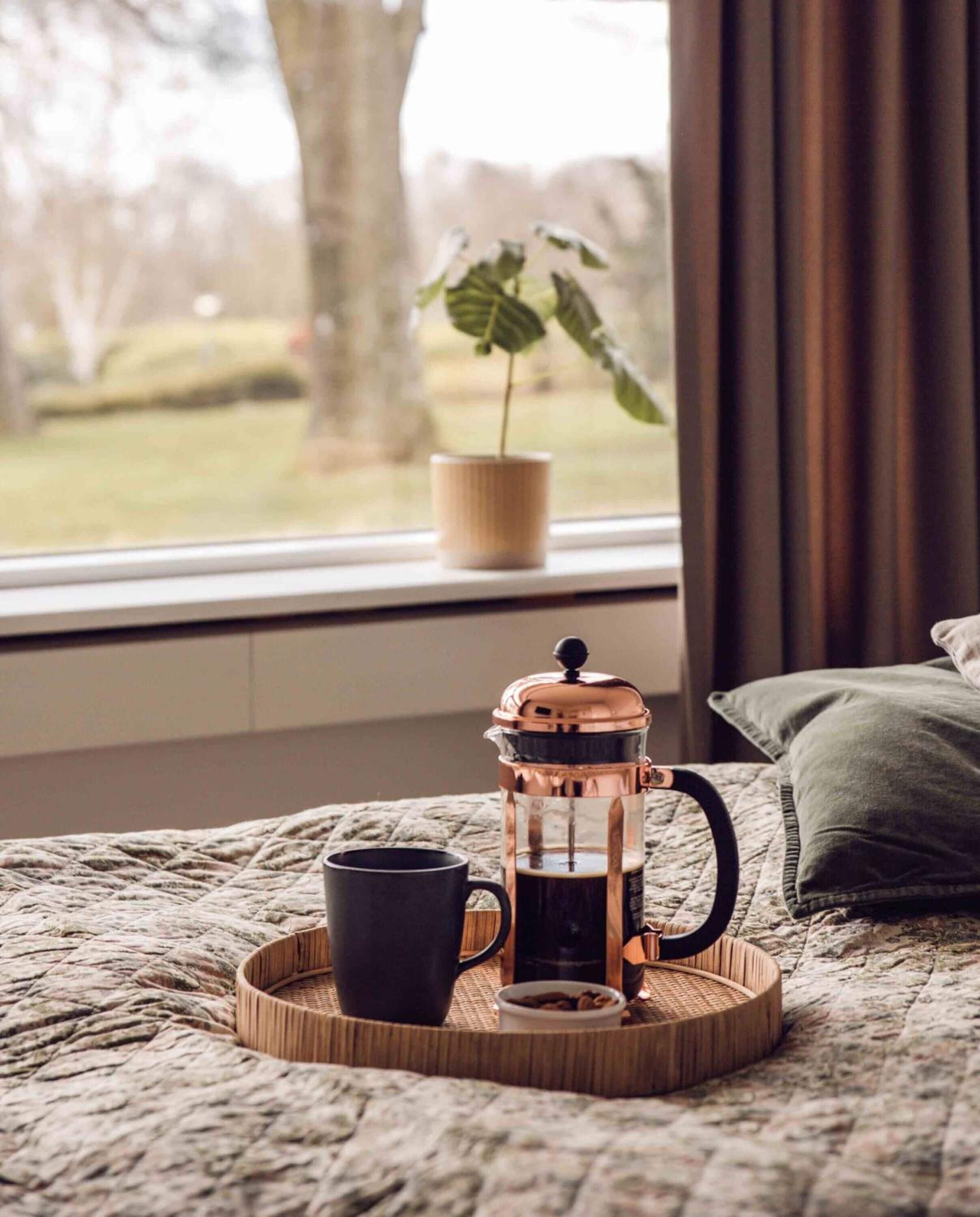 A cozy bedroom with a bed, a bedside table, a coffee maker, a cup, and a window with a view of a tree and grass.