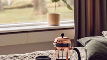 A cozy bedroom with a bed, a bedside table, a coffee maker, a cup, and a window with a view of a tree and grass.