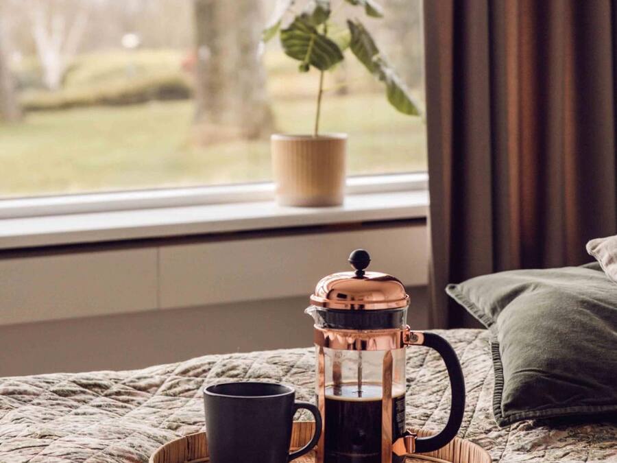 A cozy bedroom with a bed, a bedside table, a coffee maker, a cup, and a window with a view of a tree and grass.
