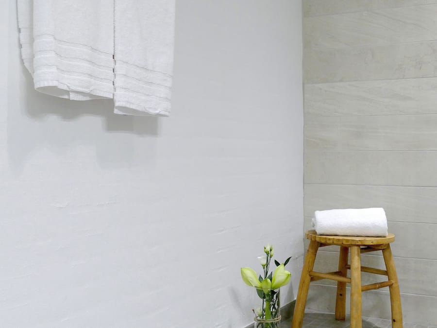 A bathroom with a white tiled floor, a wooden stool with a white towel, and a vase with green flowers on the floor.