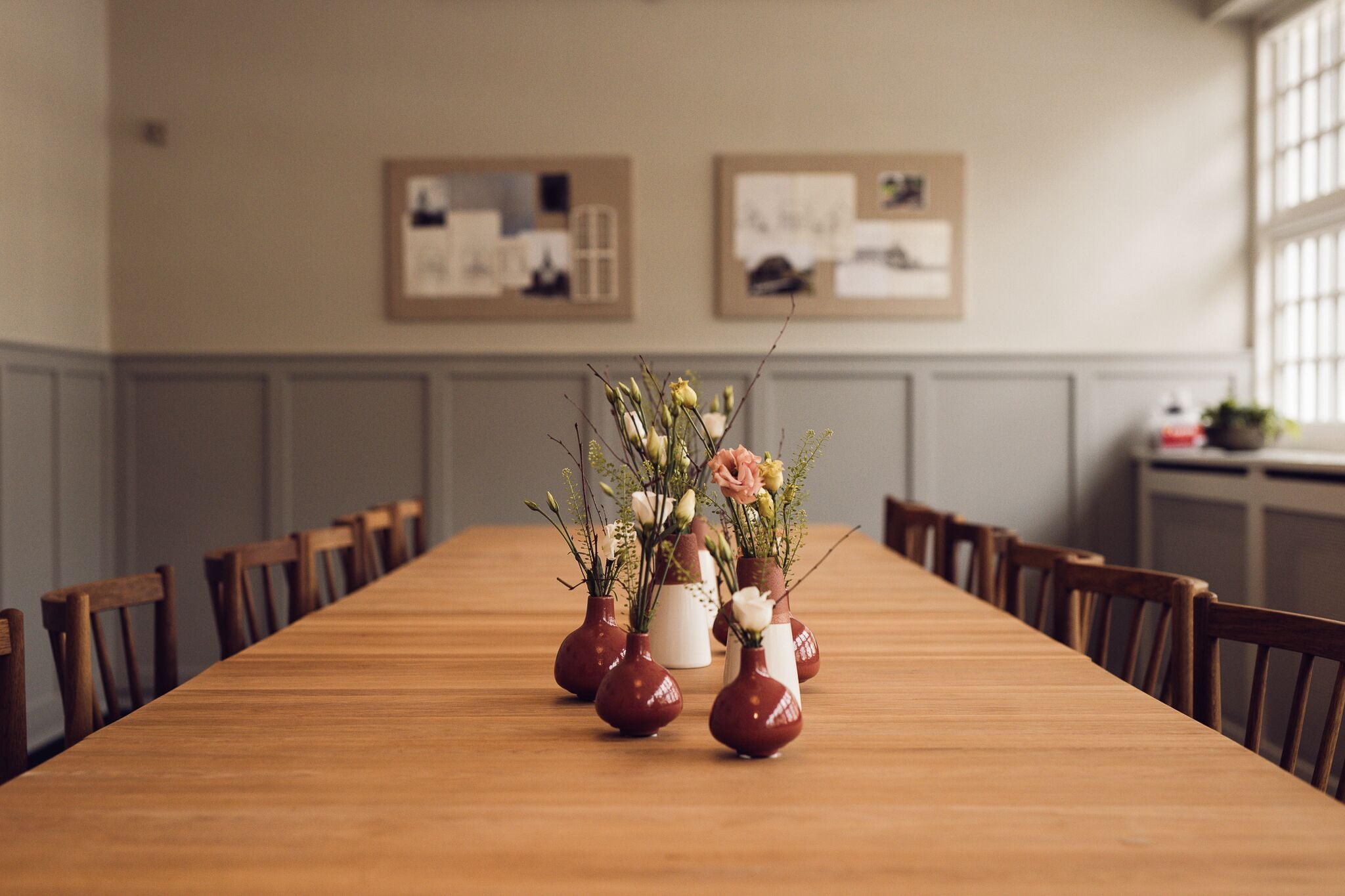 Una larga mesa de madera con arreglos florales en jarrones, sillas de madera y cuadros en la pared.