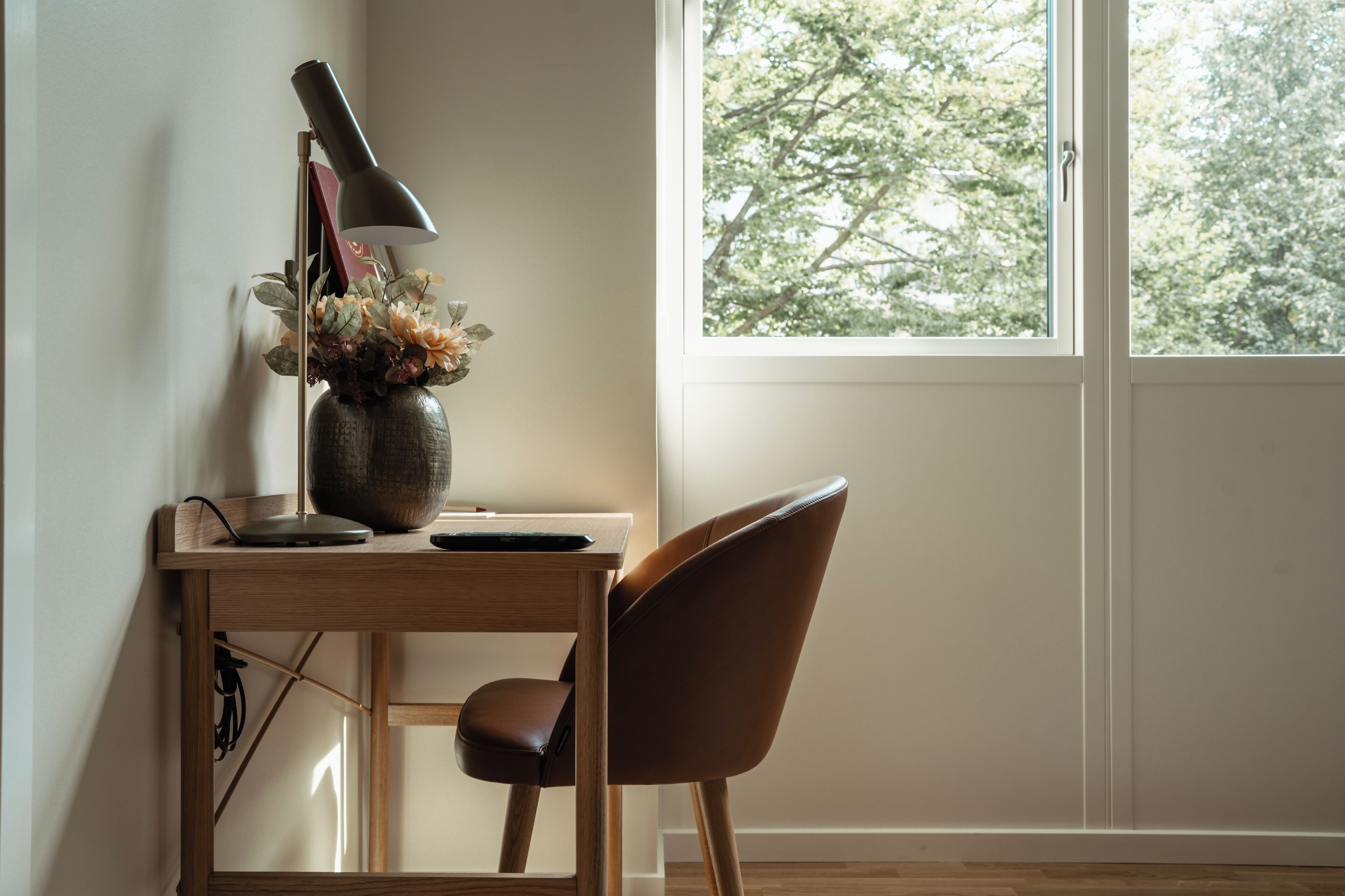 Un escritorio con una lámpara, un florero con flores y una silla en una habitación con ventana.