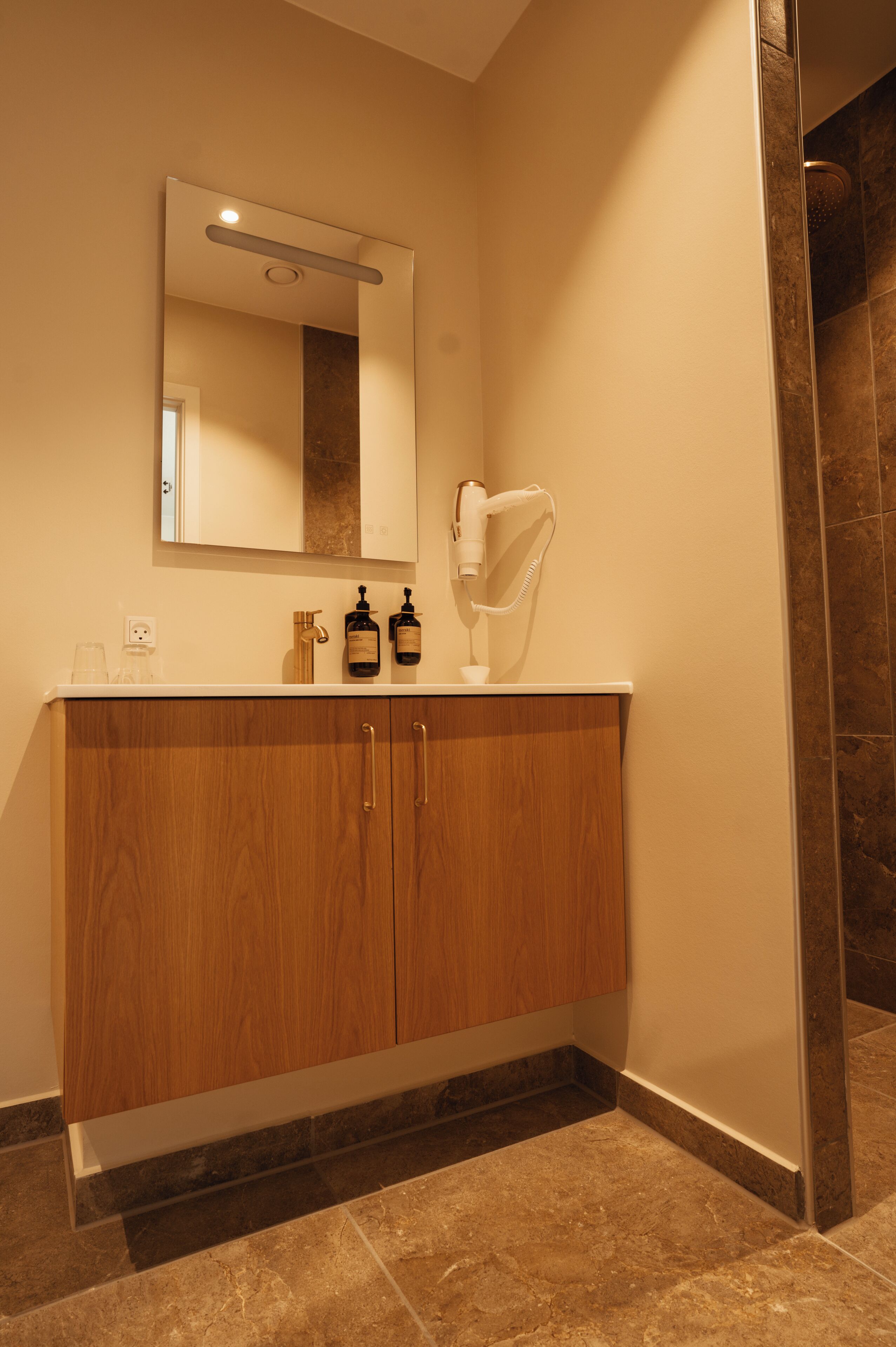 A bathroom with a wooden vanity, a mirror, a hairdryer, and a shower area.