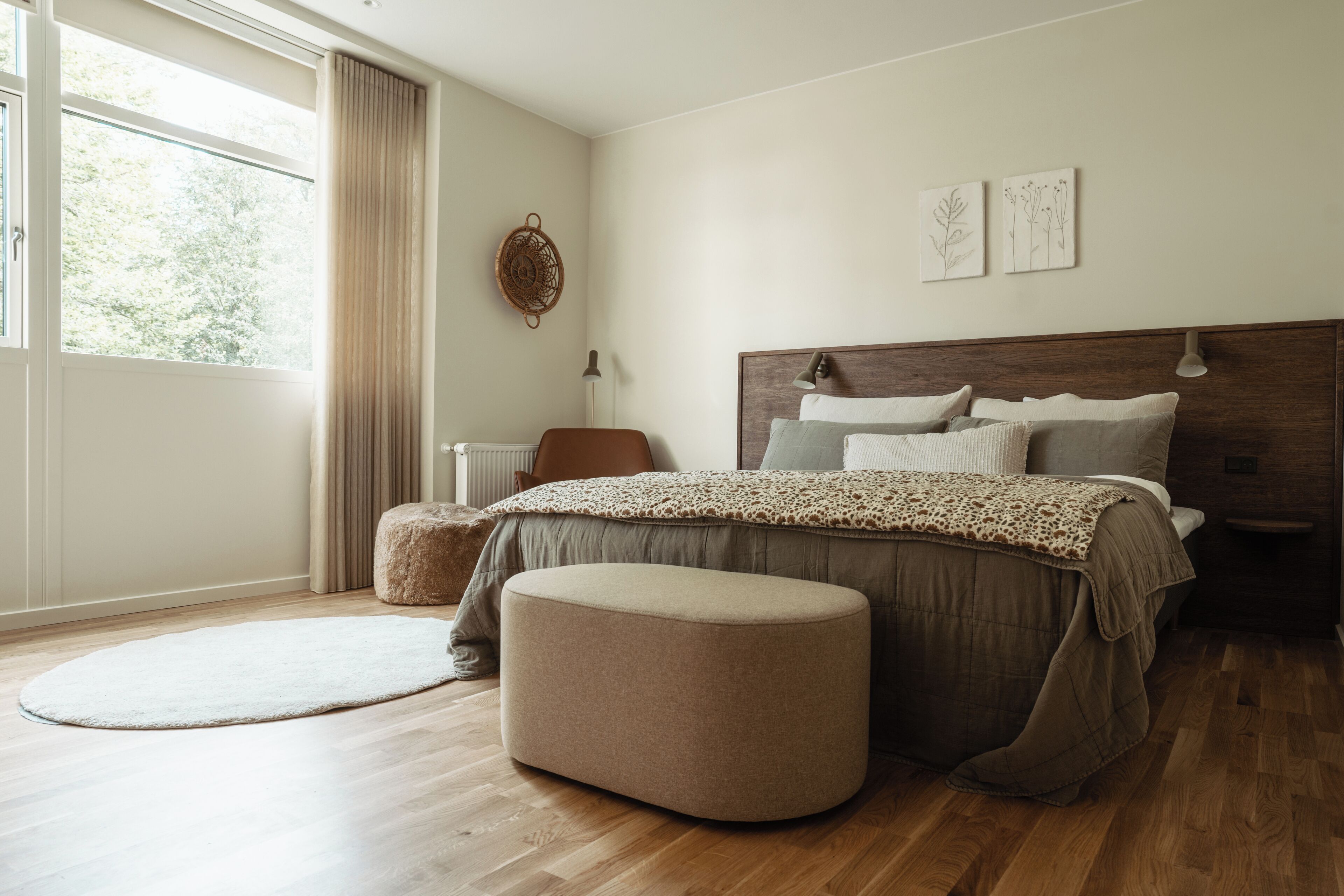 Un dormitorio con una cama grande, un reposapiés redondo, una ventana con cortinas y obras de arte en la pared.
