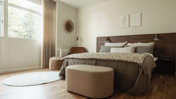 Un dormitorio con una cama grande, un reposapiés redondo, una ventana con cortinas y obras de arte en la pared.