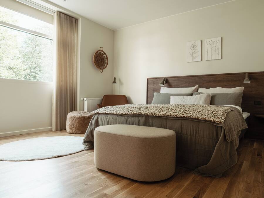 Un dormitorio con una cama grande, un reposapiés redondo, una ventana con cortinas y obras de arte en la pared.