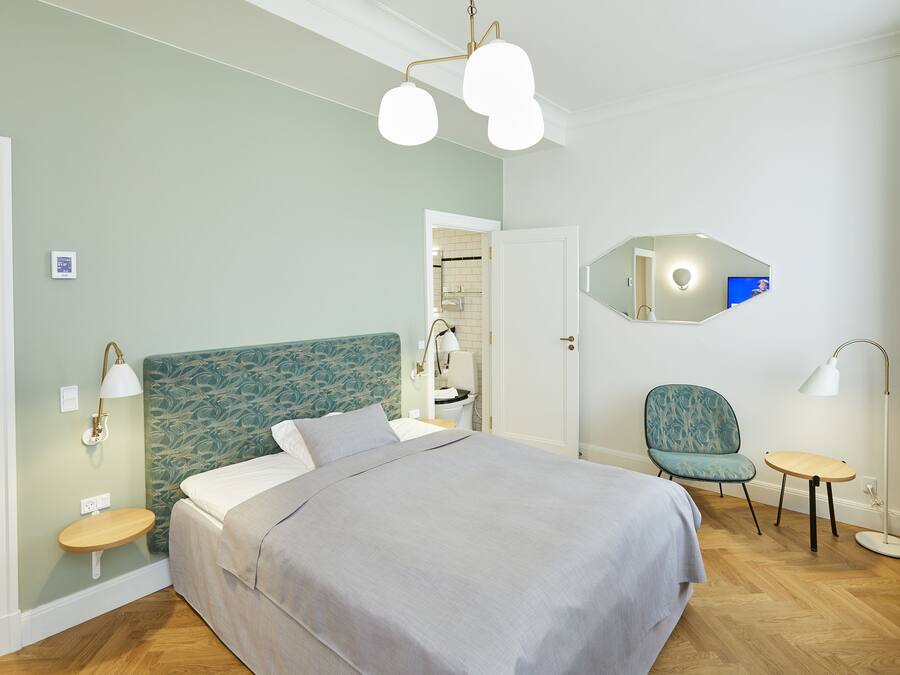 Un dormitorio con cama, una silla, una mesita, un espejo y una lámpara.