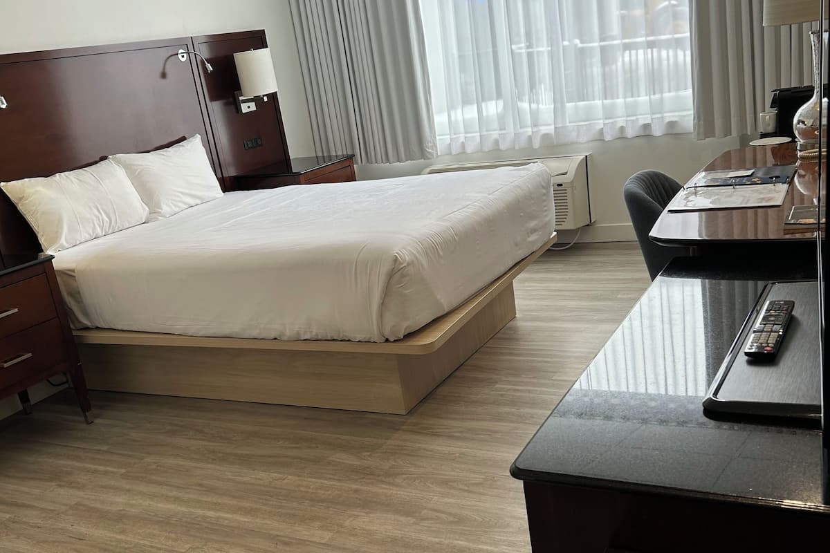 A hotel room with a large bed, a desk with a computer, and a window with blinds.
