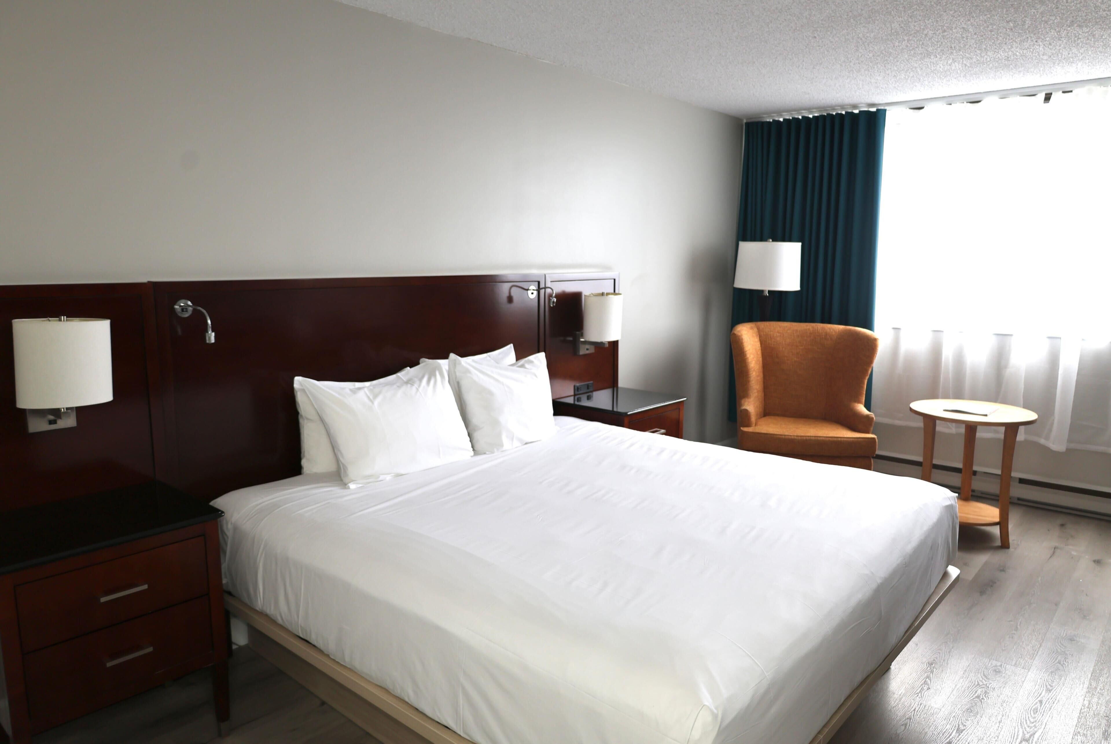 A hotel room with a bed, a nightstand, a chair, a small table, and a window with curtains.