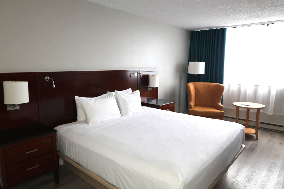 Una habitación de hotel con una cama, una silla, un escritorio, una lámpara y un ventanal con cortinas.