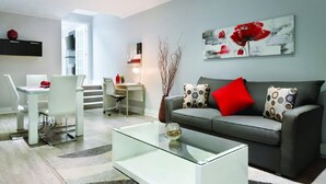 A modern living room with a glass coffee table, grey sofa with red and patterned cushions, a white dining area with chairs, and a painting on the wall. - Travelodge by Wyndham Hotel & Convention Centre Quebec City (Québec City)